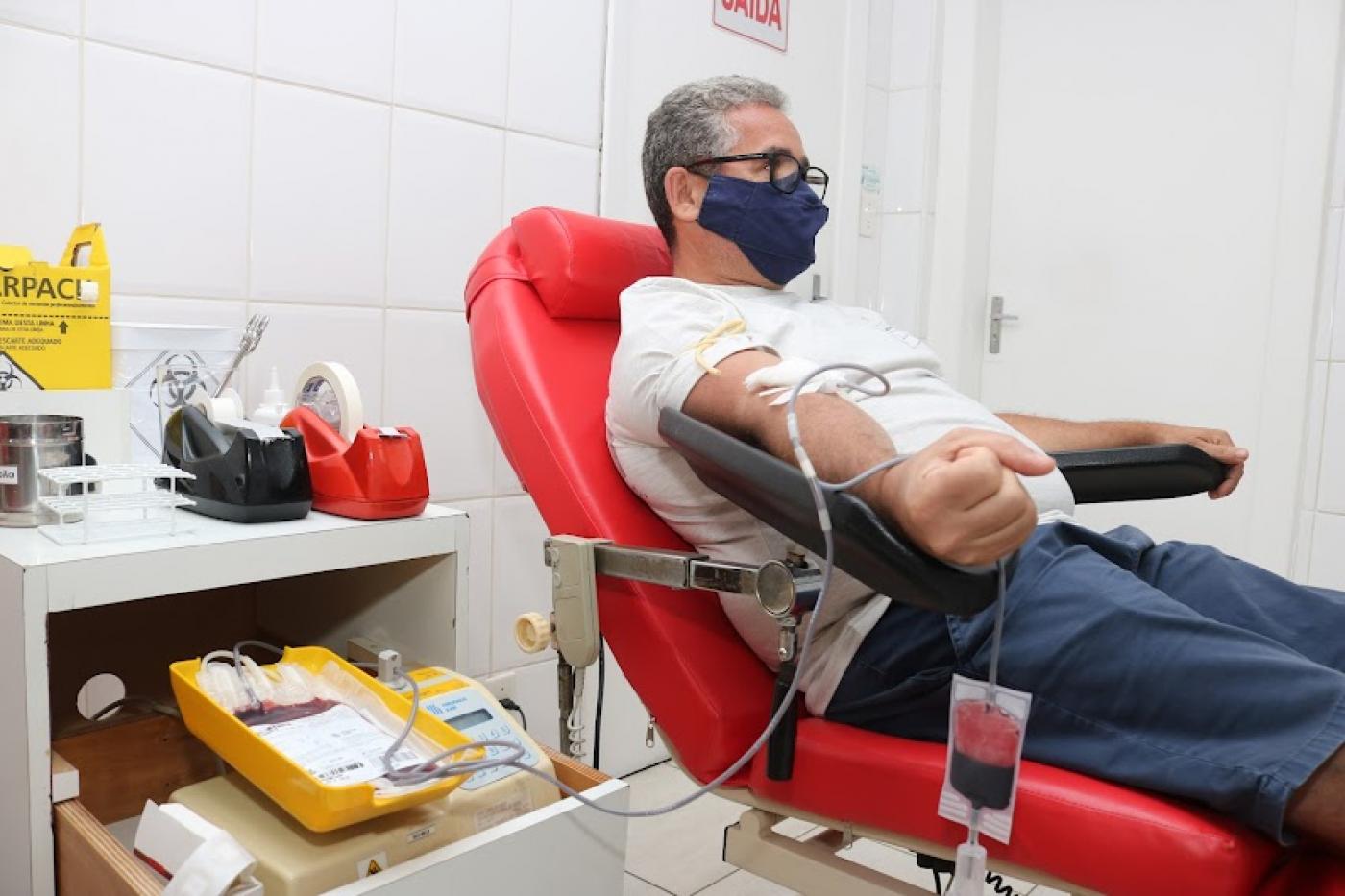 homem esta sentado em cadeira com apetrechos para doação de sangue. A bolsa já com quantidade do líquido está pendurada na cadeira. #paratodosverem