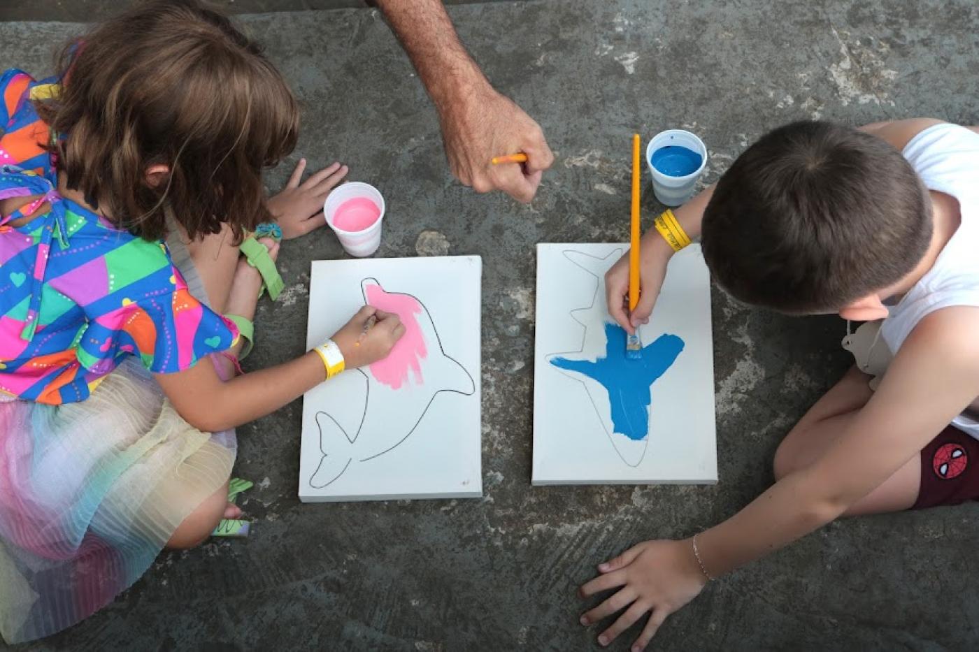 foto tirada do alto mostra duas crianças frente a frente pintando telas. A silhueta de animais marinhos está pronta. Elas pintam sobre a área delimitada.  #paratodosverem 