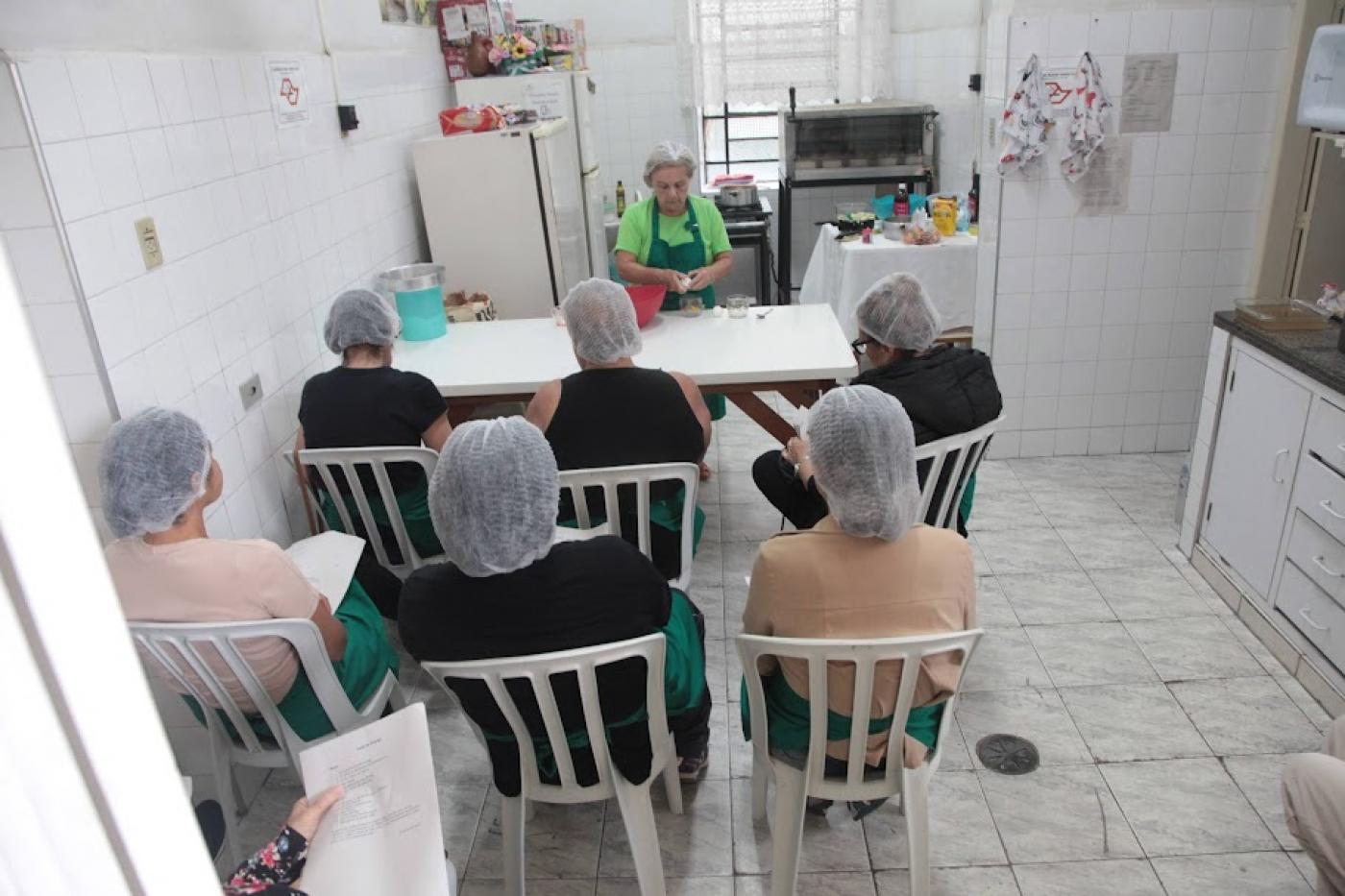 mulheres sentadas de costas para a foto, todas usando tocas, prestam atenção em aula de culinária dada por uma mulher à frente de uma mesa. #paratodosverem 