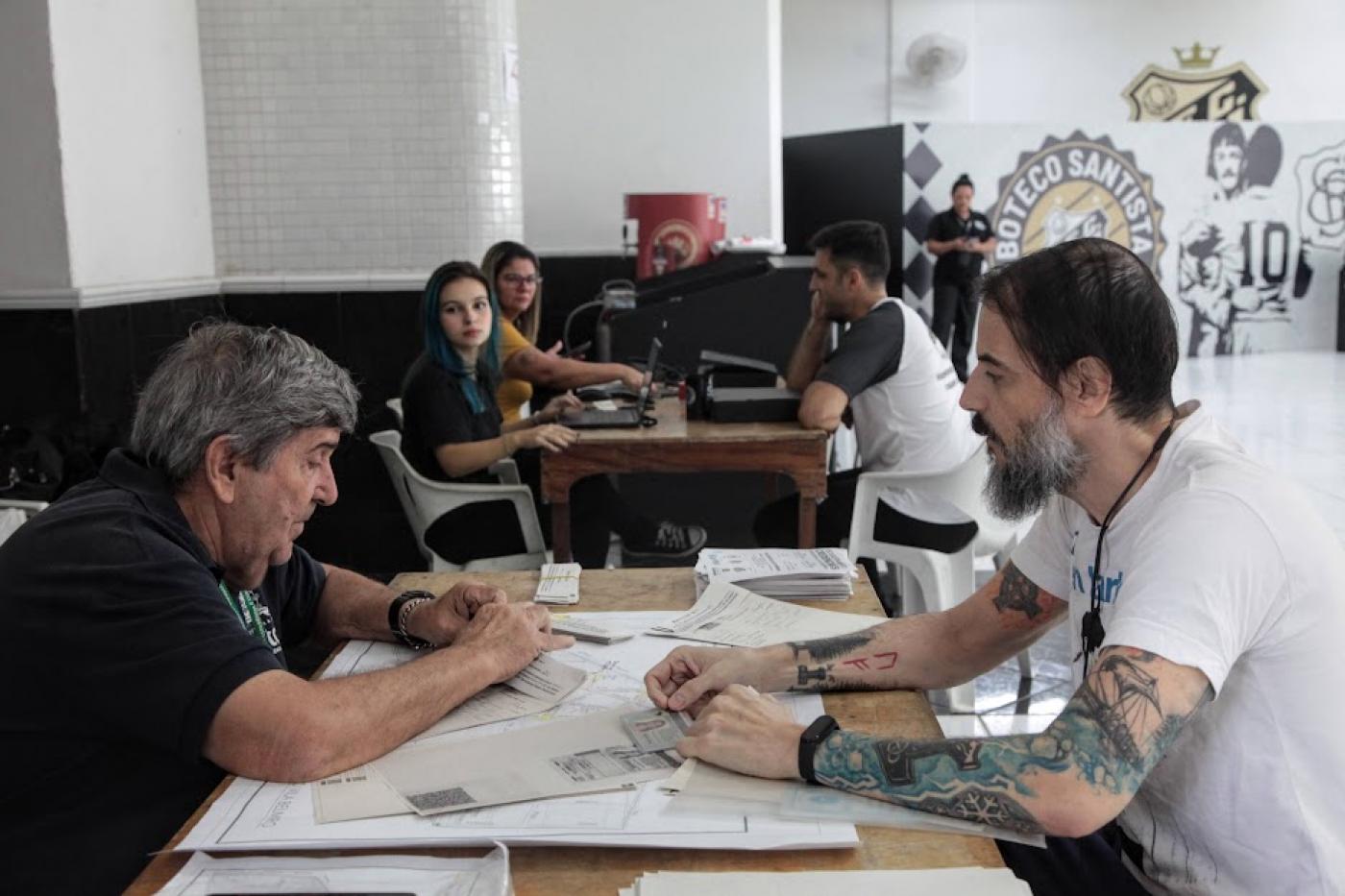 dois homens sentados frente a frente com uma mesa no meio. Um deles entrega documentos. #paratodosverem