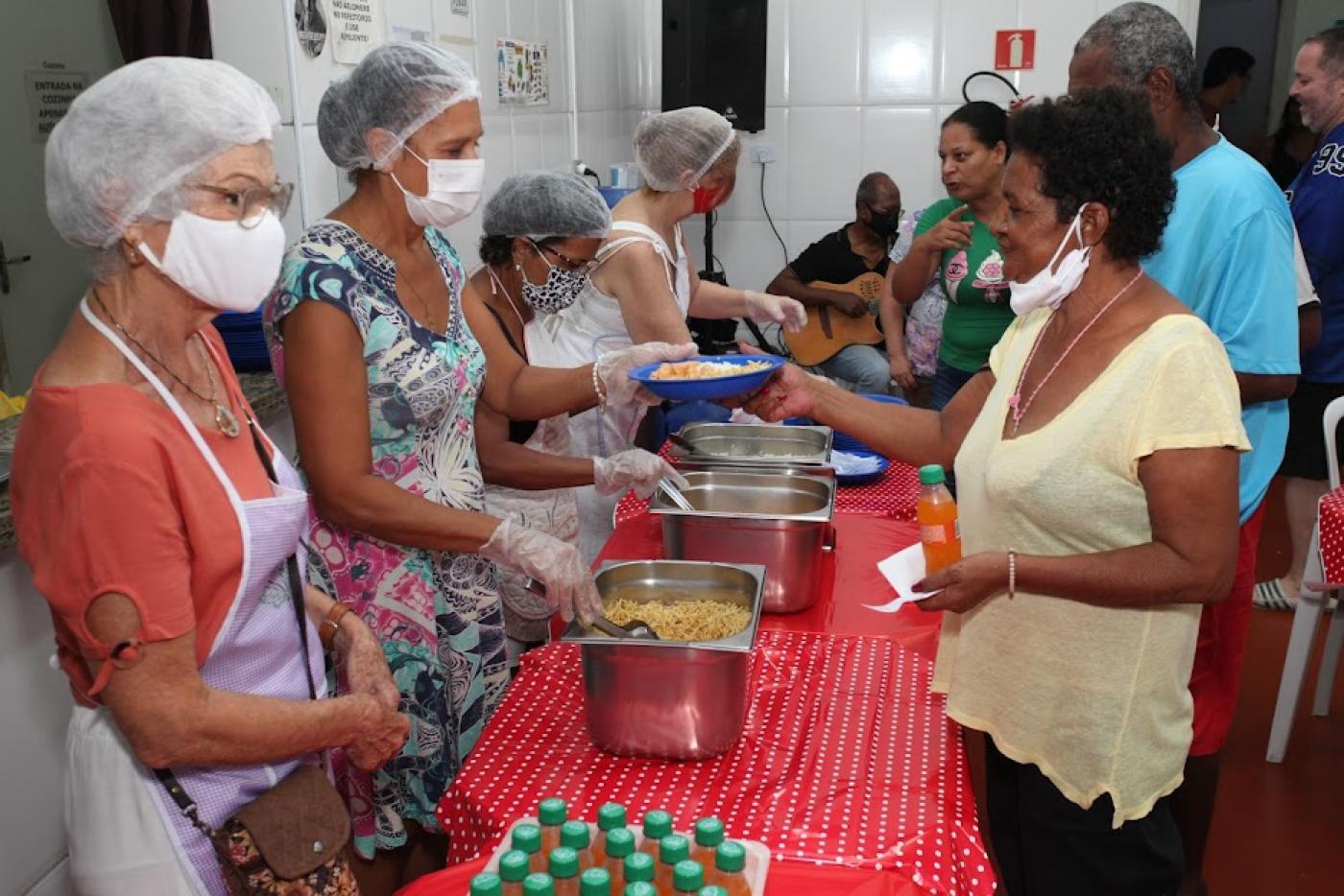 mesa com travessas fundas com comida. De um lado da mesa, pessoas servem. Do outro, são servidas. #paratodosverem