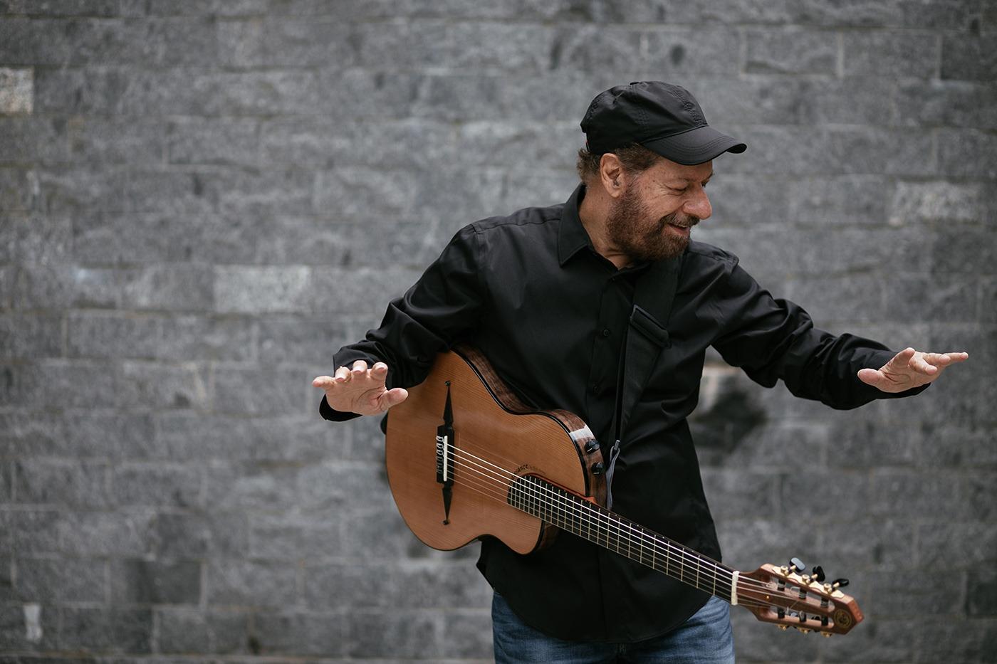 cantor está com violão pendurado ao lado do corpo. As mãos estão no ar. ele canta. #paratodosverem 