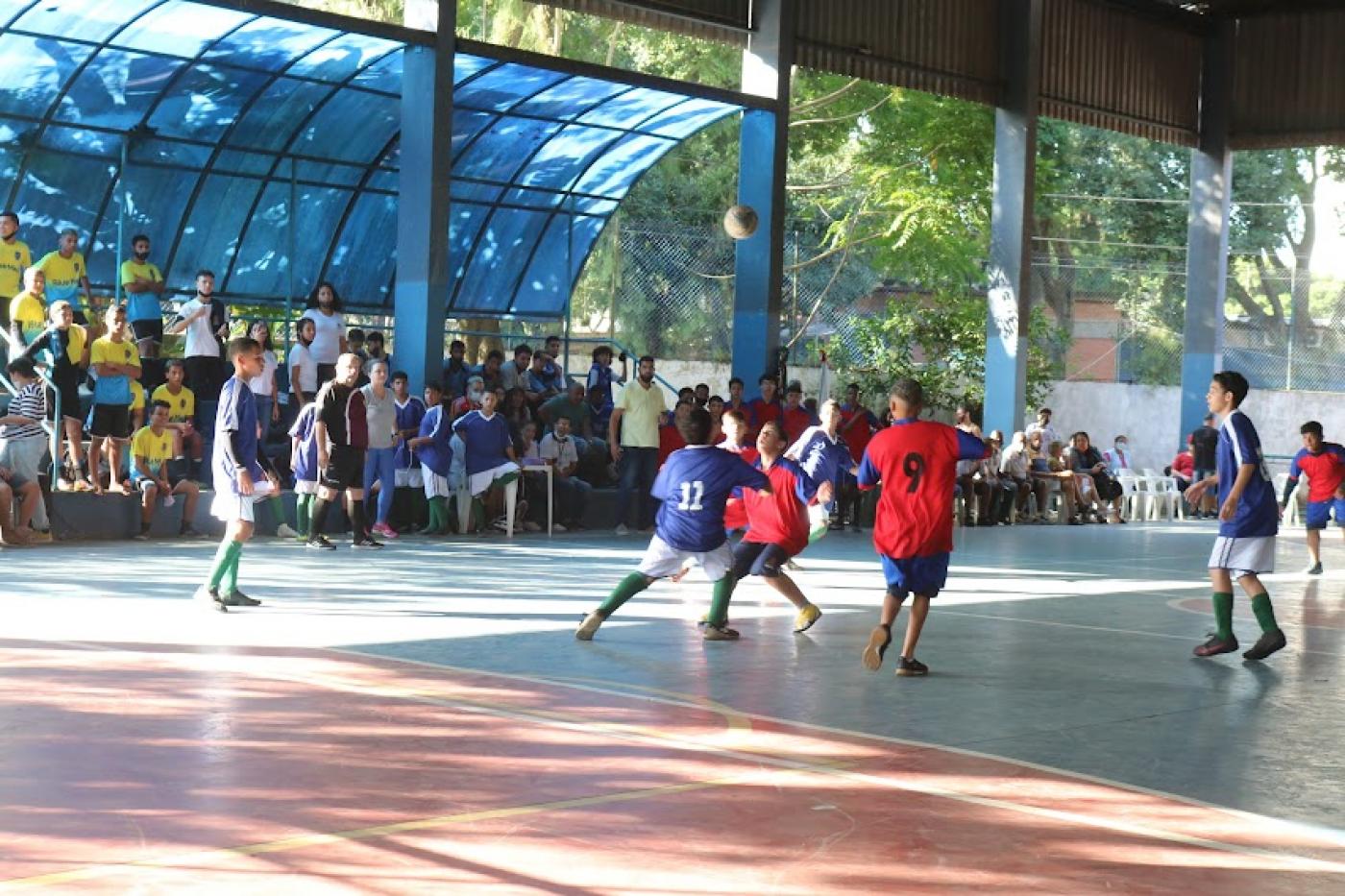 crianças estão jogando futebol em pátio. Ao fundo e e à esquerda o público está na arquibancada. #paratodosverem