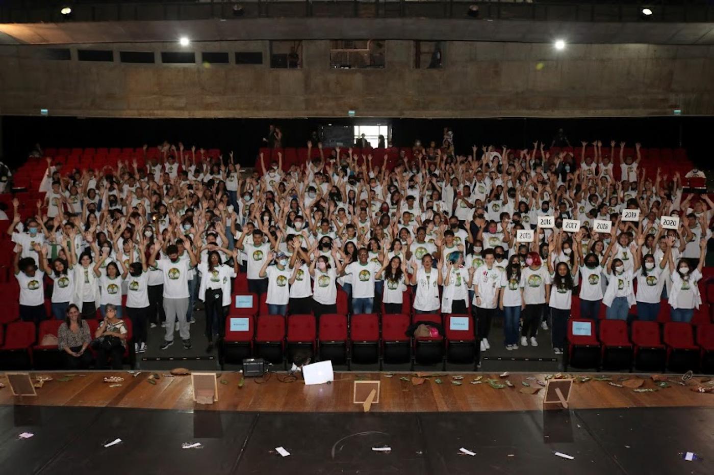 centenas de jovens uniformizados e em pé com as  mãos para o alto na plateia de teatro. #paratodosverem