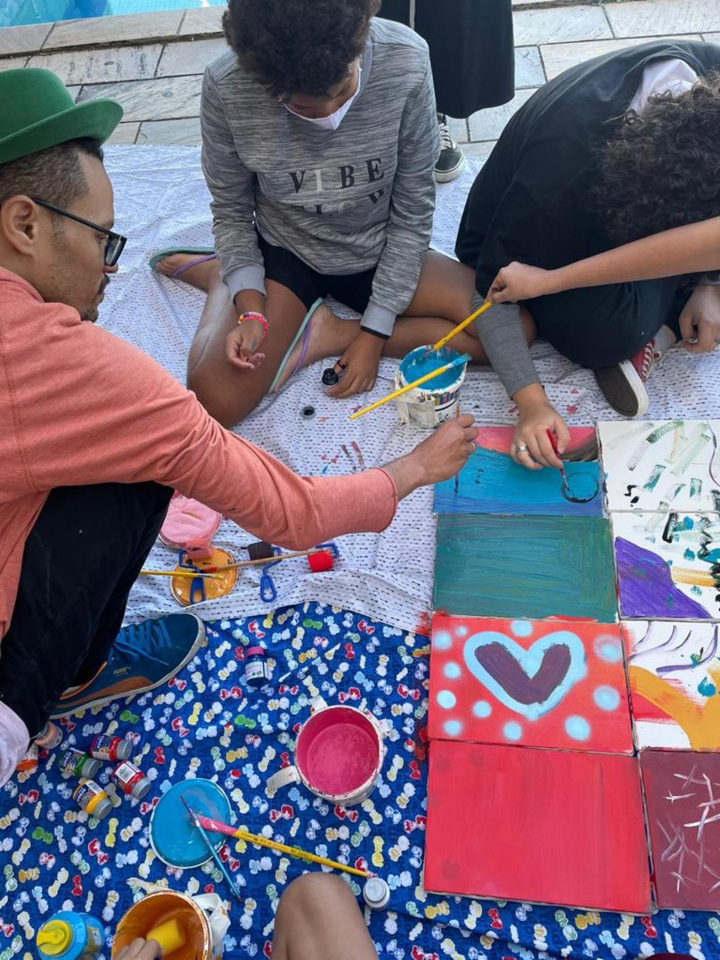 jovens estão sentados no chão, com pinceis. Eles pintam sobre tecidos. #paratodosverem