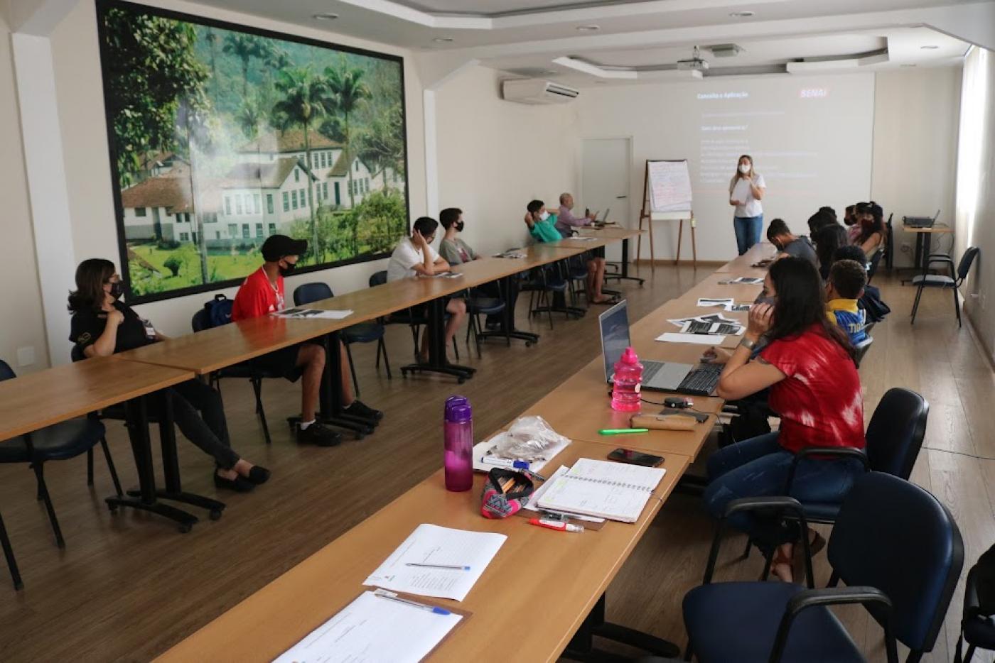 Jovens estão sentados à mesas colocadas lado a lado. Eles olham para a mulher que está ao fundo da sala, que está diante de uma projeção na parede. #paratodosverem