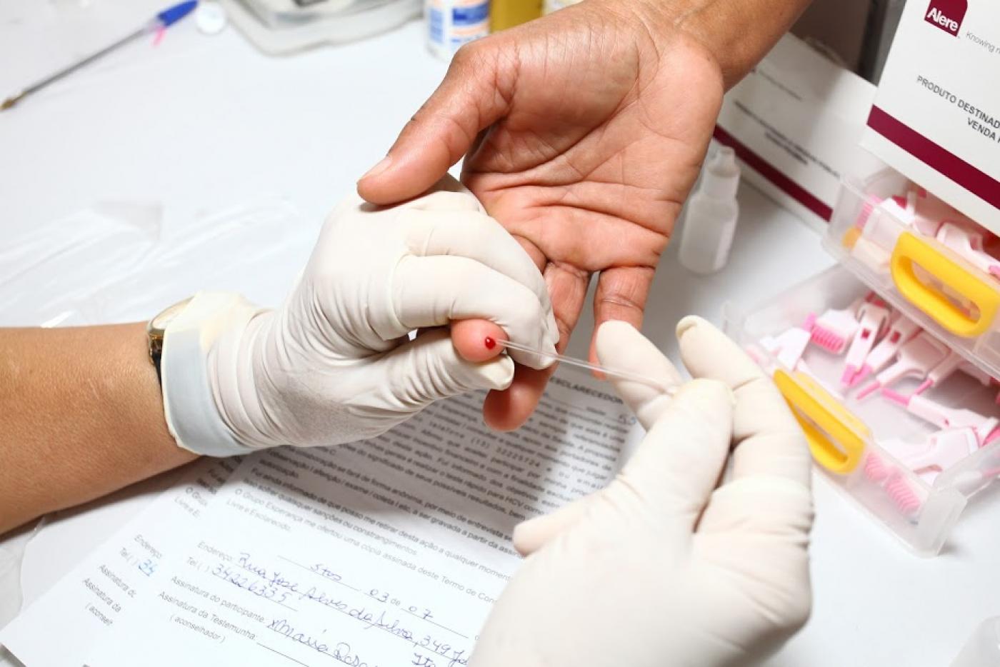 mão com luva segura fura a ponta do dedo de uma pessoa para extrair uma gota de sangue. #paratodosverem