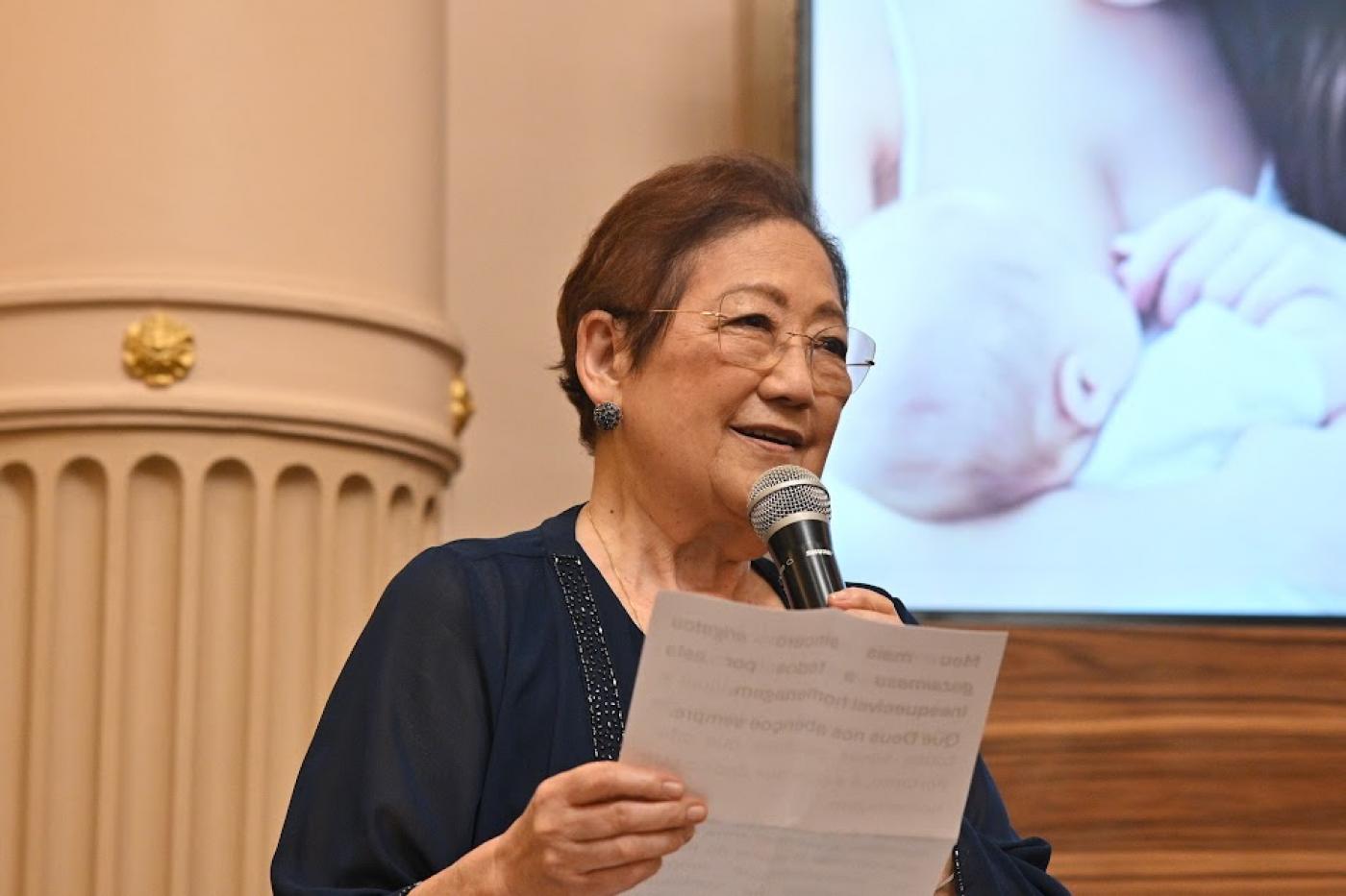 a médica lê discurso. ao fundo, em tela, mulher amamenta bebê. #paratodosverem