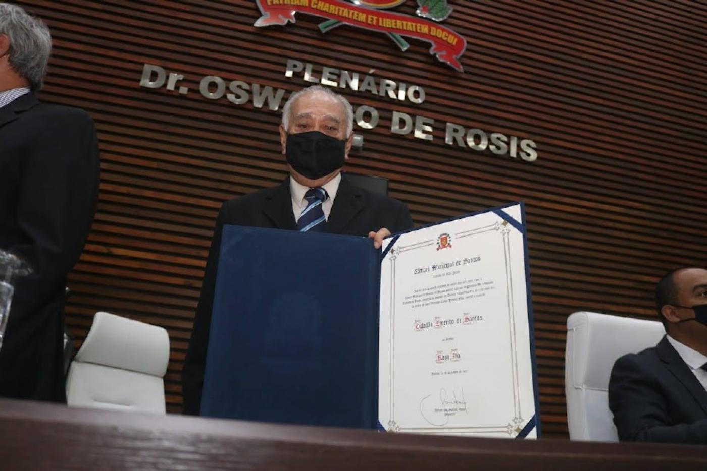 o homenageado posa para foto usando máscara e segurando o diploma concedido pela câmara. #paratodosverem