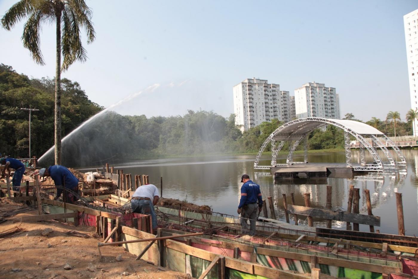 Vista geral dá área em torno da lagoa, com homens trabalhando no piso, o palco no meio da água e mais fundo, prédios do morro. #Paratodosverem