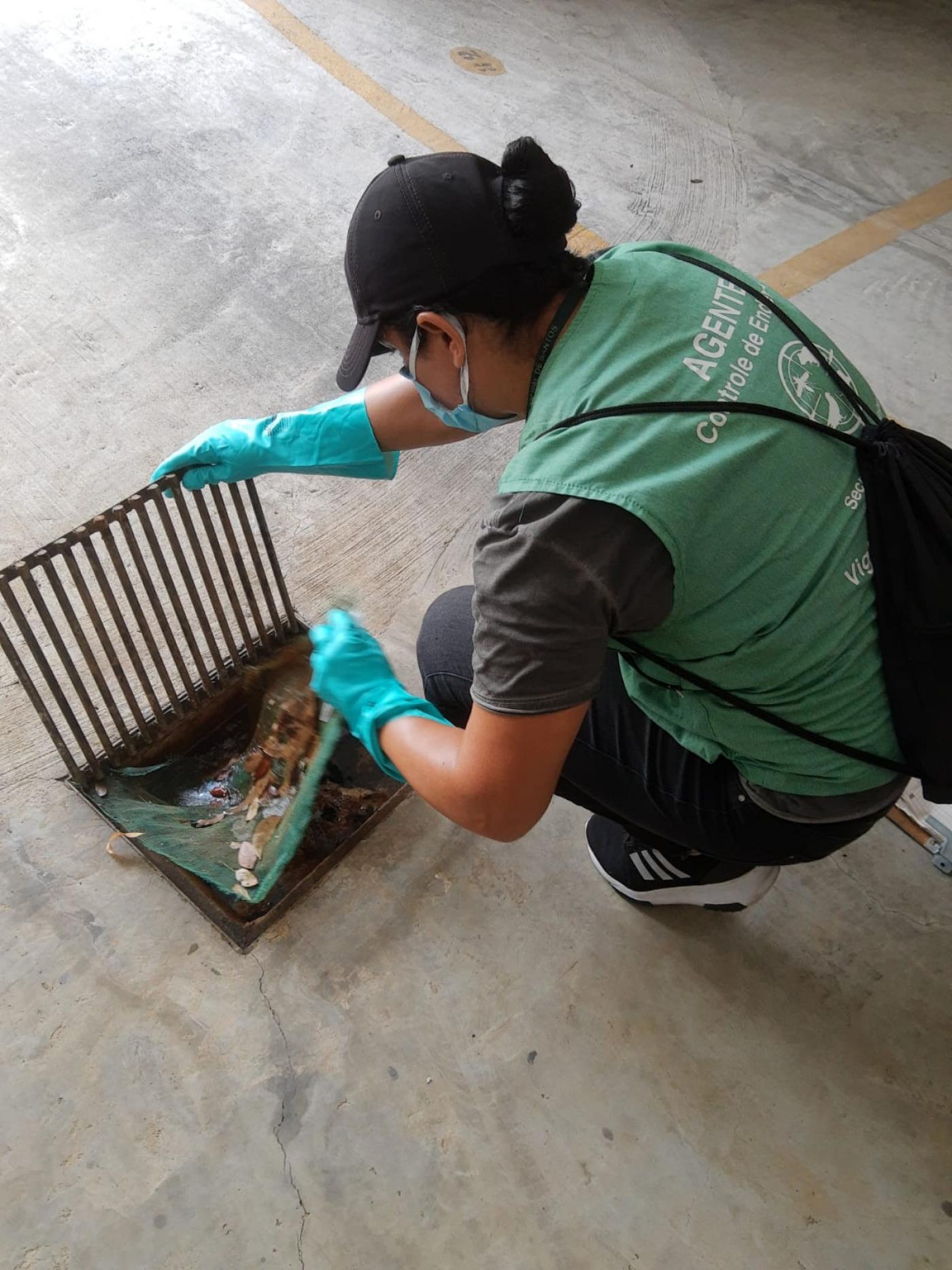 Mulher identifica por colete está agachada junto a um ralo. Ela mexe na tela de proteção. #Paratodosverem