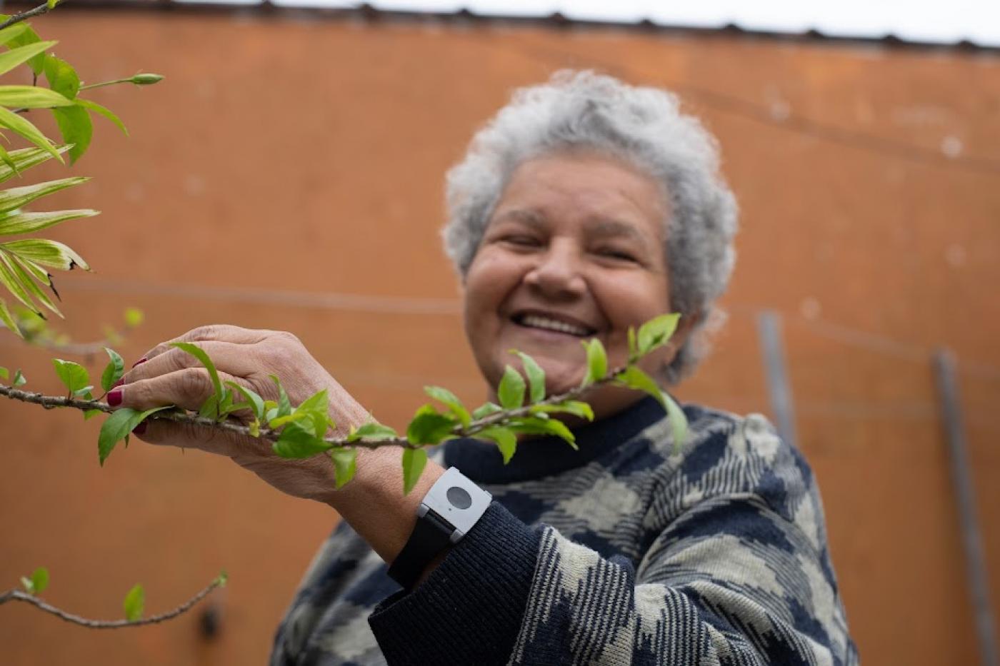 mulher está mexendo em uma planta. No braço esquerdo ela tem um aparelho, similar a um relógio. #paratodosverem