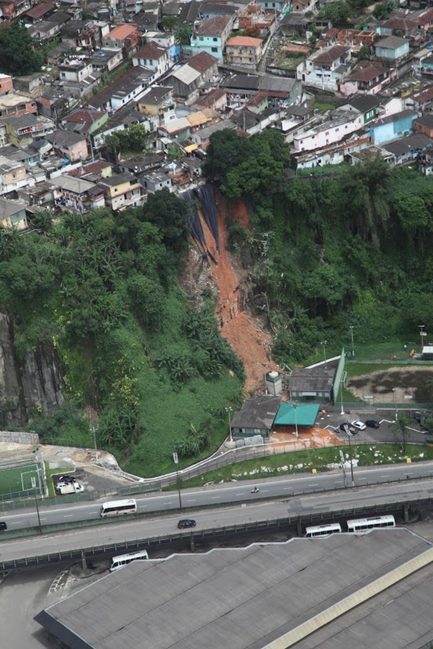 Área de morro onde houve deslizamento. Imagem feita com drone mostra o alto do morro, com moradias e uma faixa de deslizamento na encosta. Avenida passa em baixo. #Paratodosverem