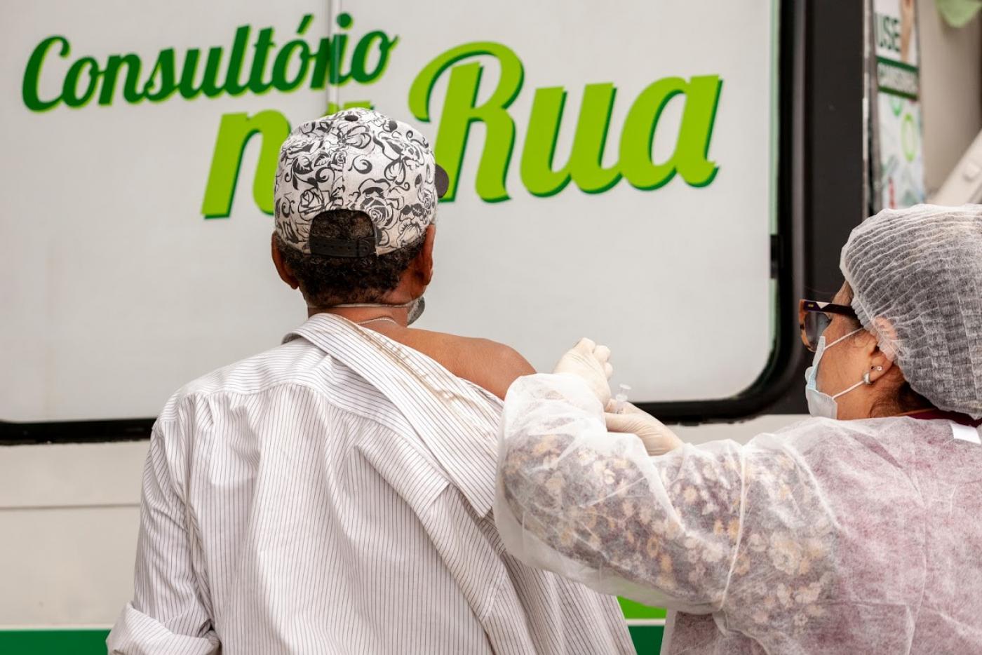 Mulher usando avental, gorro, luva e máscara vacina homem que está de costas. Ele usa boné. Ao fundo se lê:Consultório na Rua impresso na lateral de uma van. #Paratodosverem