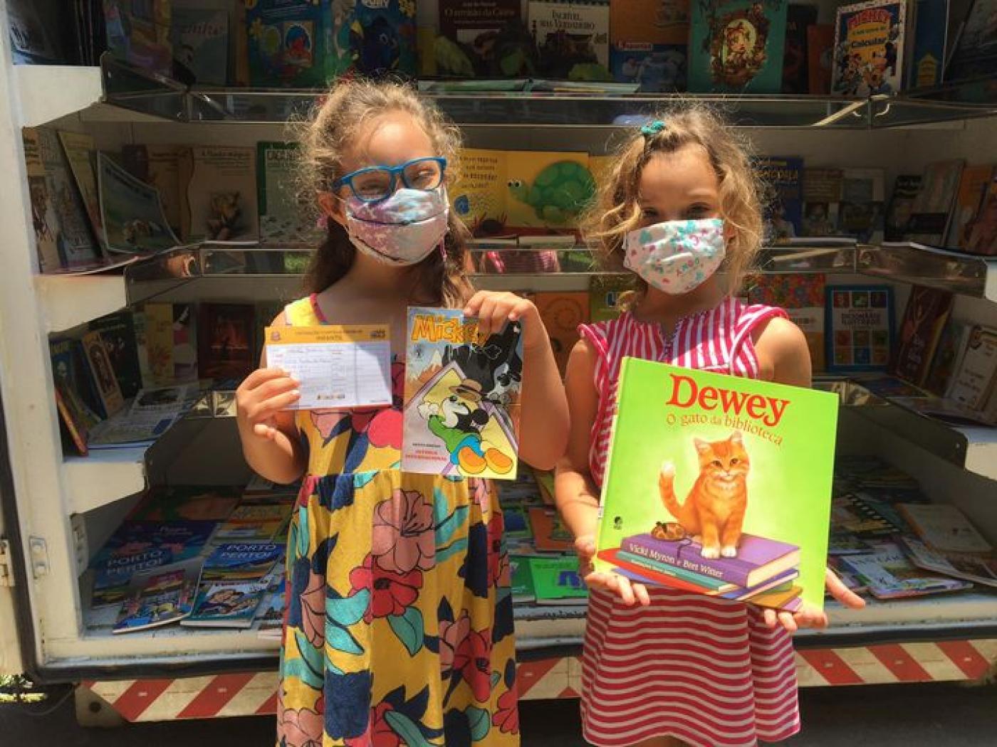 duas meninas seguram livros em frente a perua que funciona como biblioteca. #paratodosverem