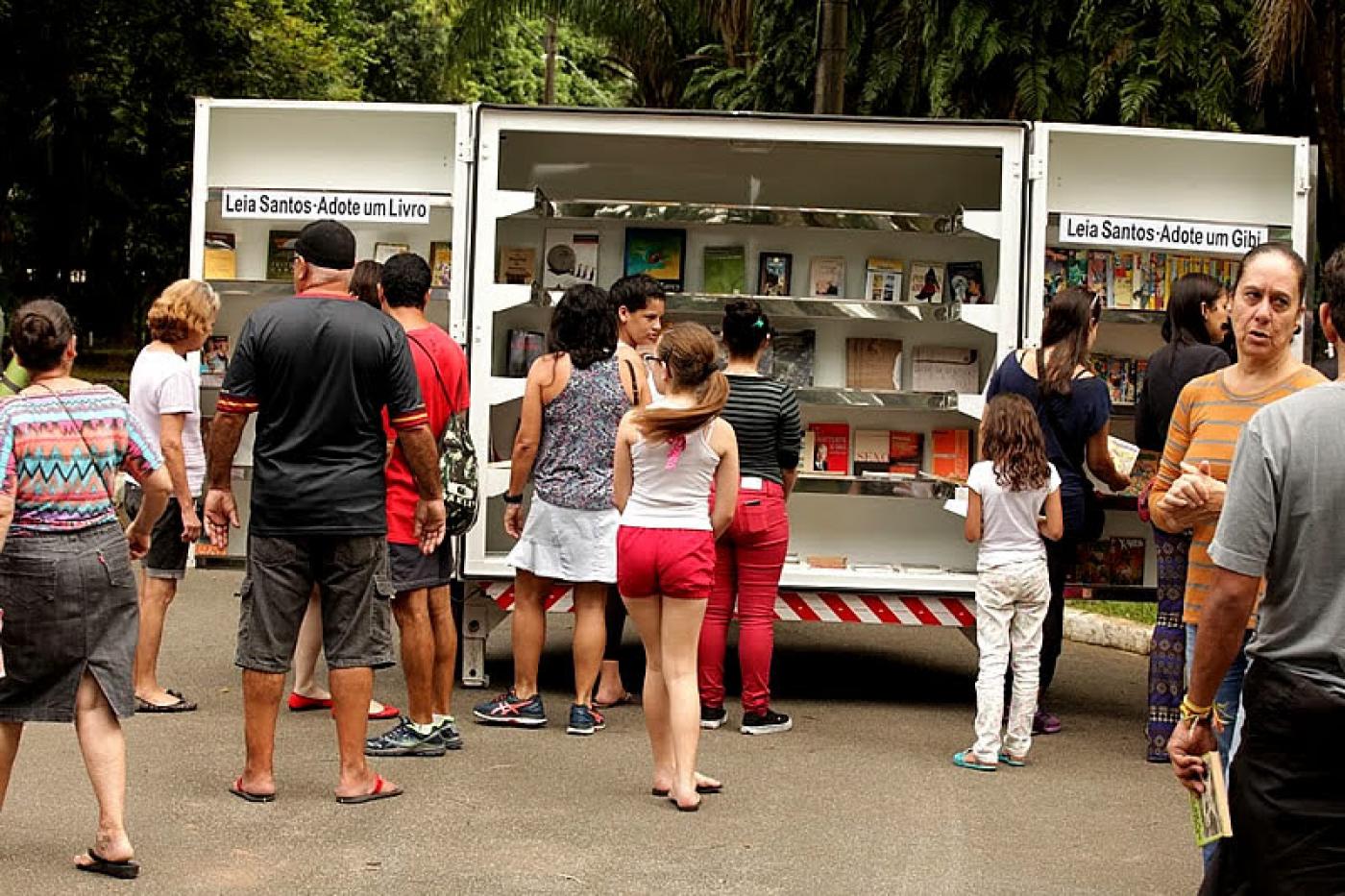 Estante com vários livros aberta em rua. Várias pessoas estão em torno. #Pracegover