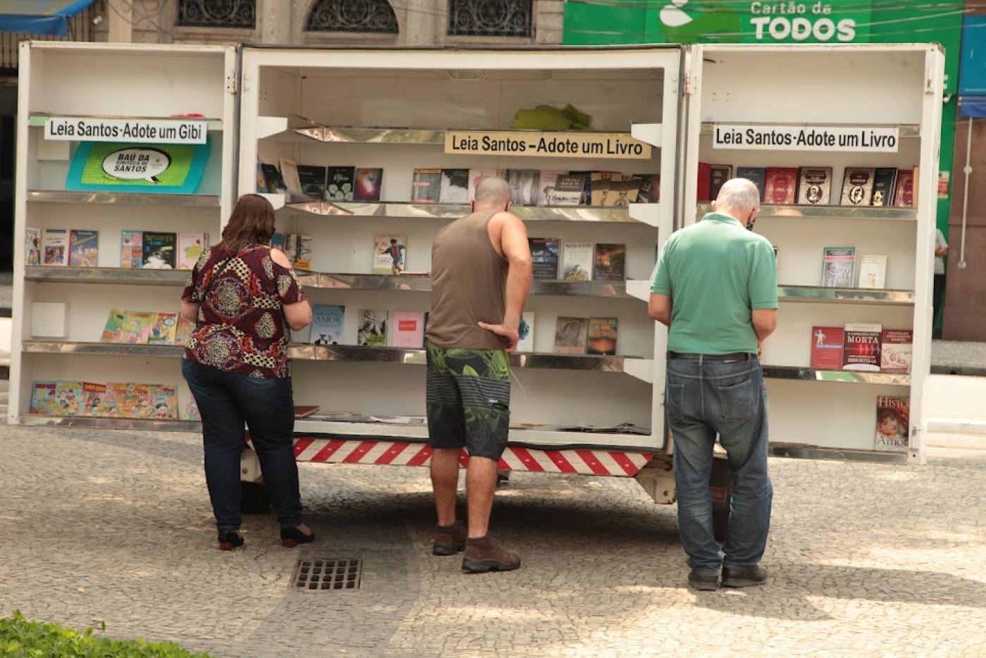 três pessoas estão diante de biblioteca ambulante. Todsos estão de costas para a foto. #paratodosverem