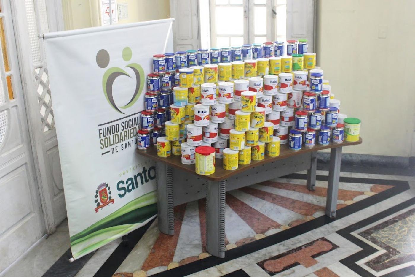 mesa com várias latas de leite em pó empilhadas. ao lado esquerdo, o banner do fundo social de solidariedade