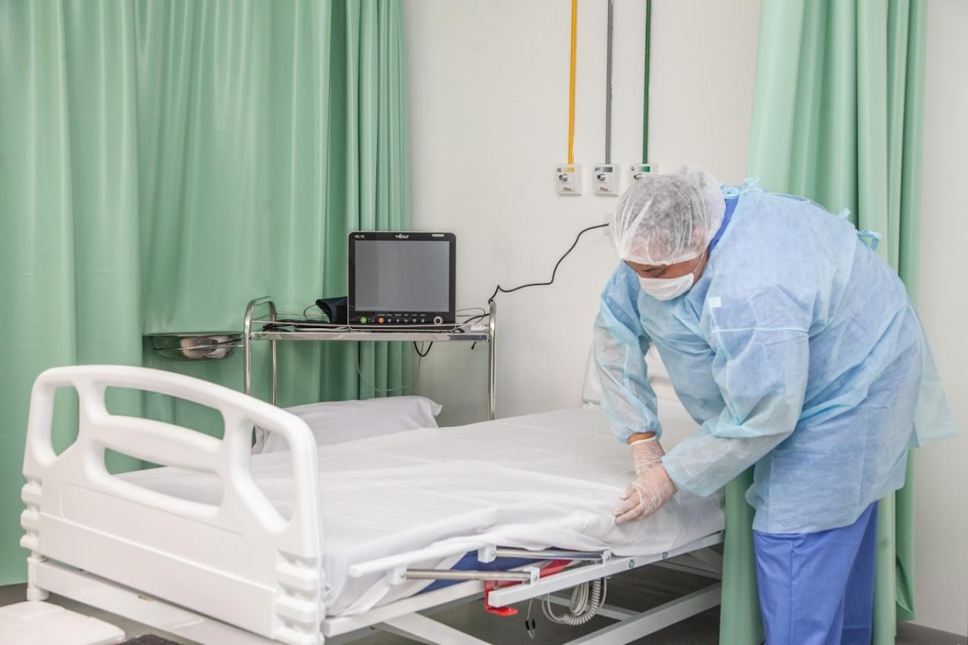 Mulher usando avental, luvas, máscara e gorro prepara leito em hospital. Ela está encaixando o lençol no colchão. Há canos de oxigênio fixados na parede atrás dela. Cortinas recolhidas fazem  o papel de biombo. #Paratodosverem