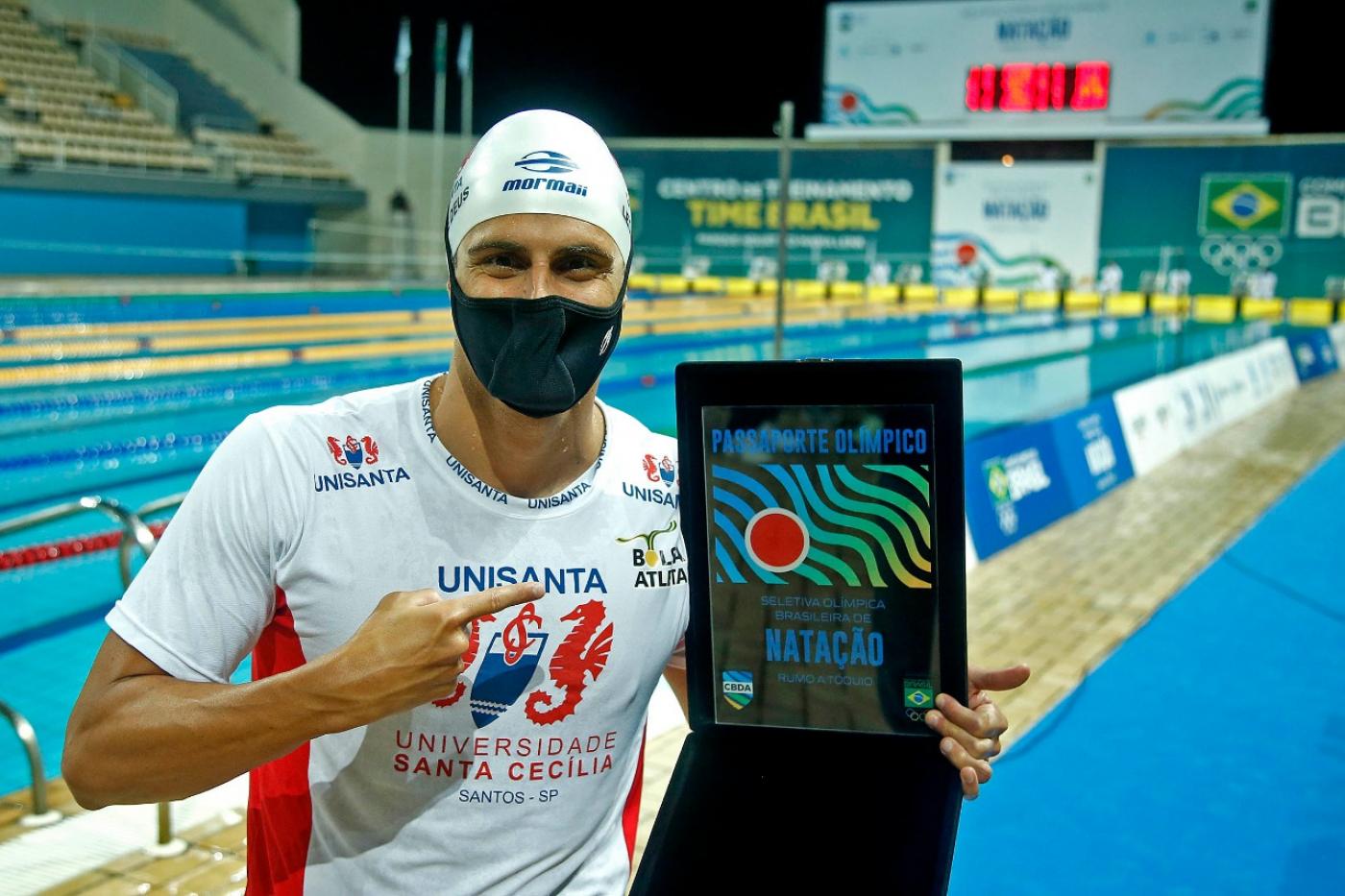 O nadador está de touca e camiseta, de costas para a piscina e segurando uma medalha que representa a classificação para a Olimpíada. #paratodosverem