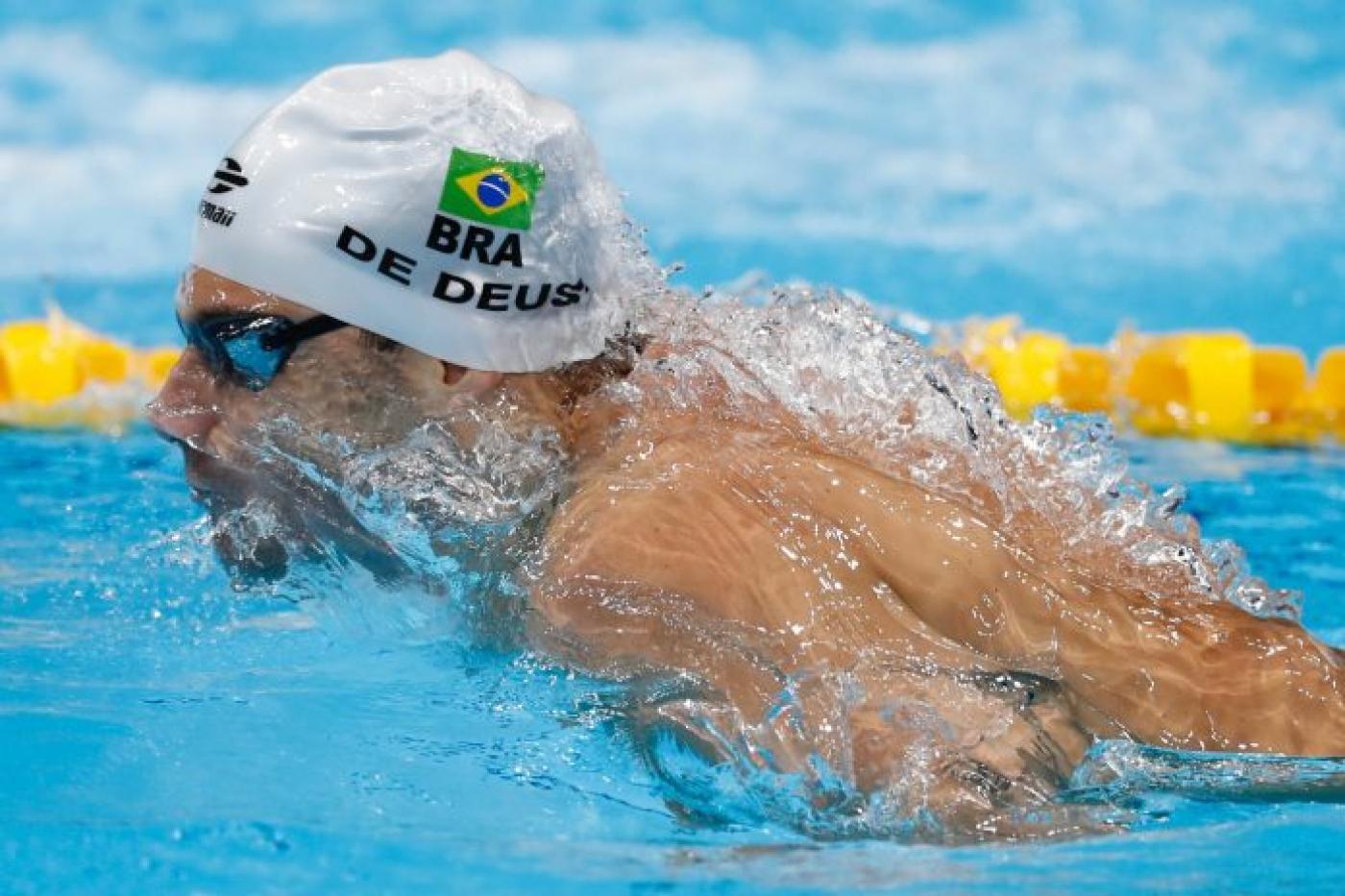 Imagem mostra o atleta nadando com toca branca com bandeira do Brasil e nome De Deus. #pracegover