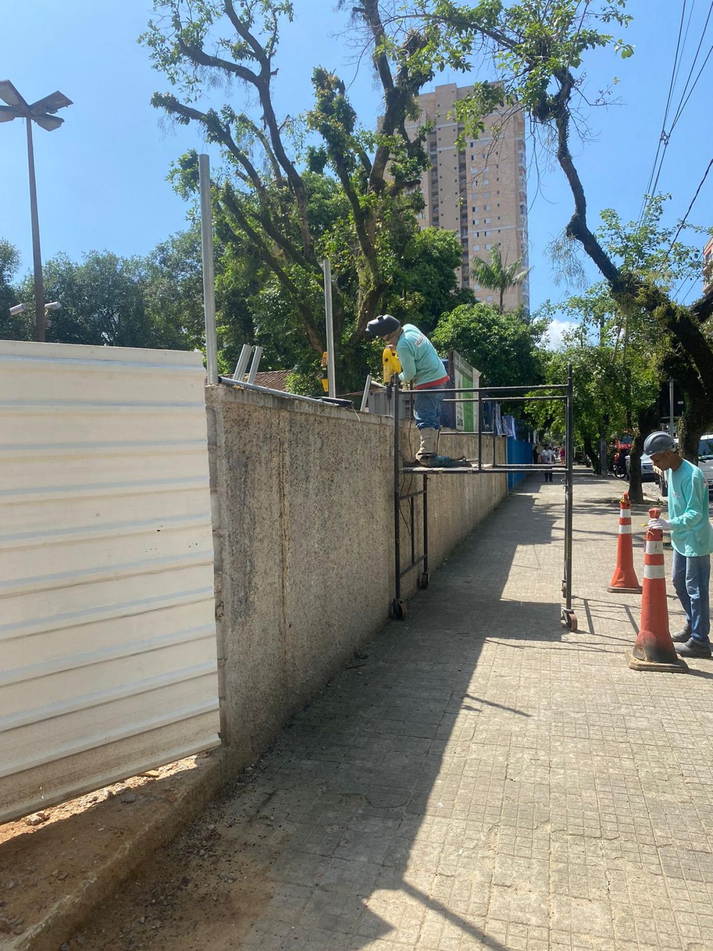homem está sobre andaime instalando alambrado em muro. Ele está do lado de fora, na calçada. #paratodosverem 