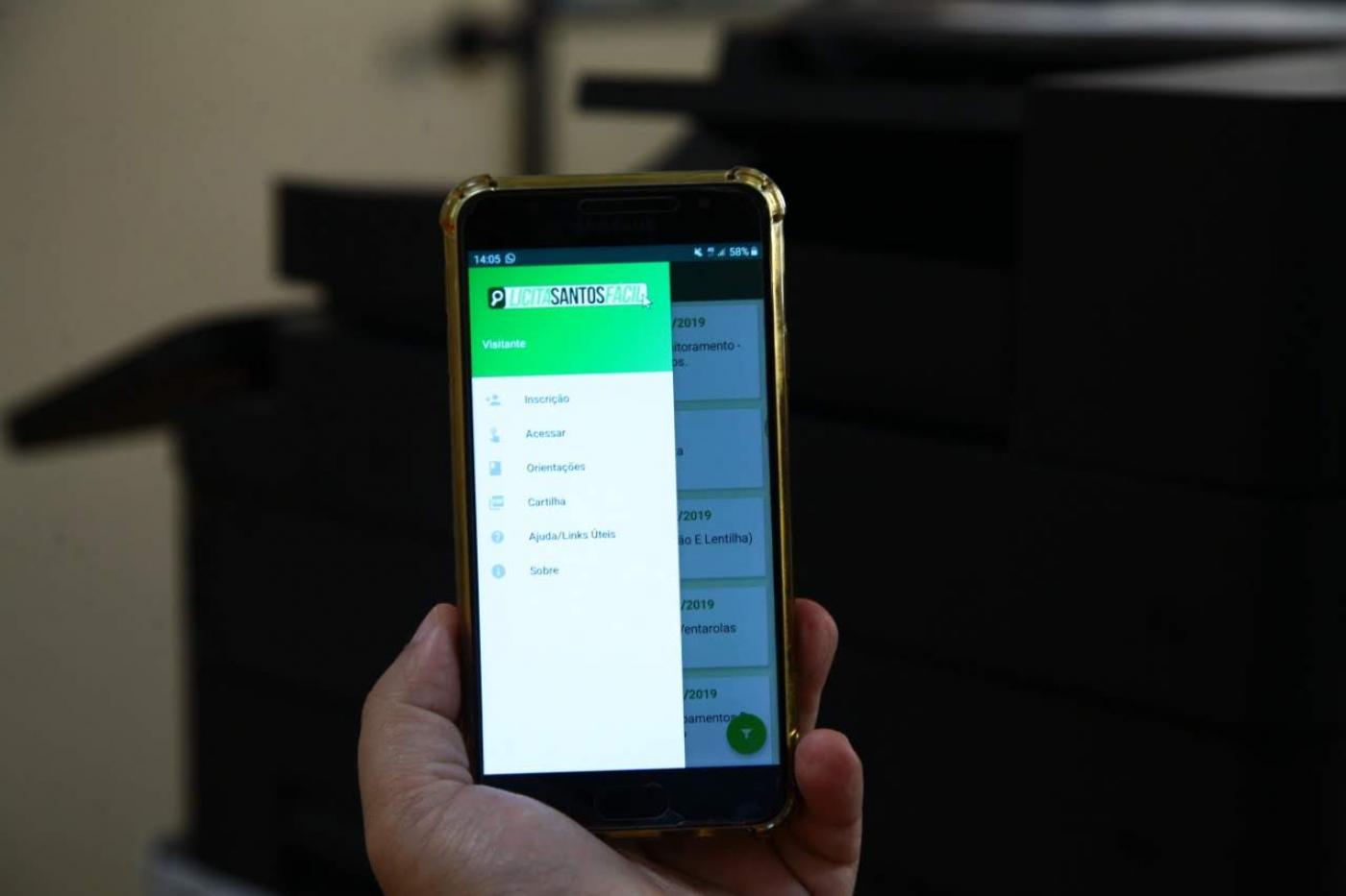 Mão está segurando smartphone com tela aberta no programa Licita Santos Fácil. #Pracegover