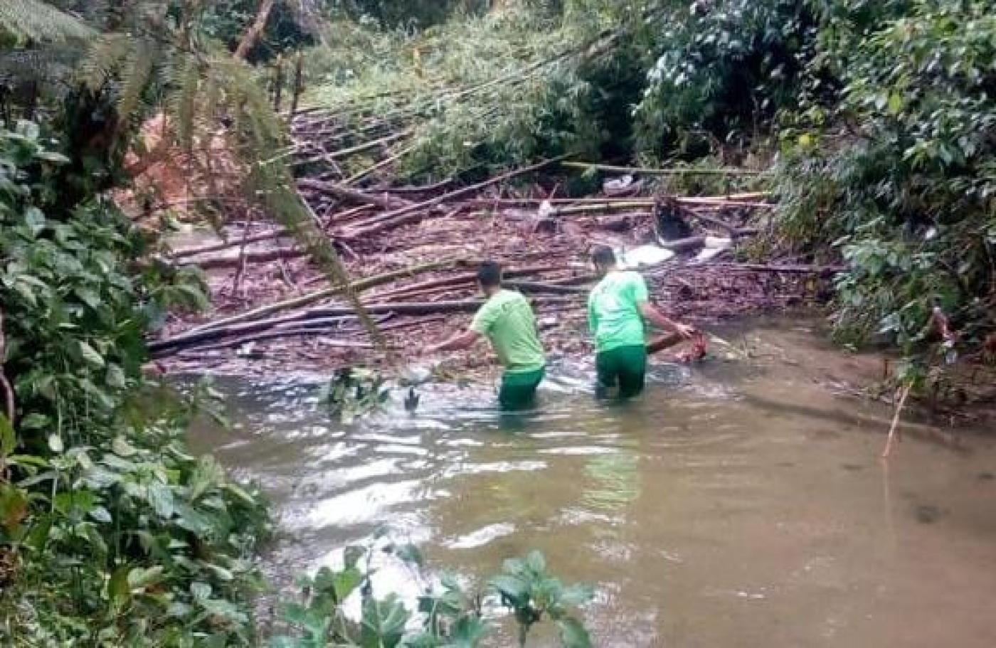 homens uniformizados estão dentro de rio com água na altura da coxa. Eles mexem em galhos que estão obstruindo trecho. Há mata nas laterais #paratodosverem