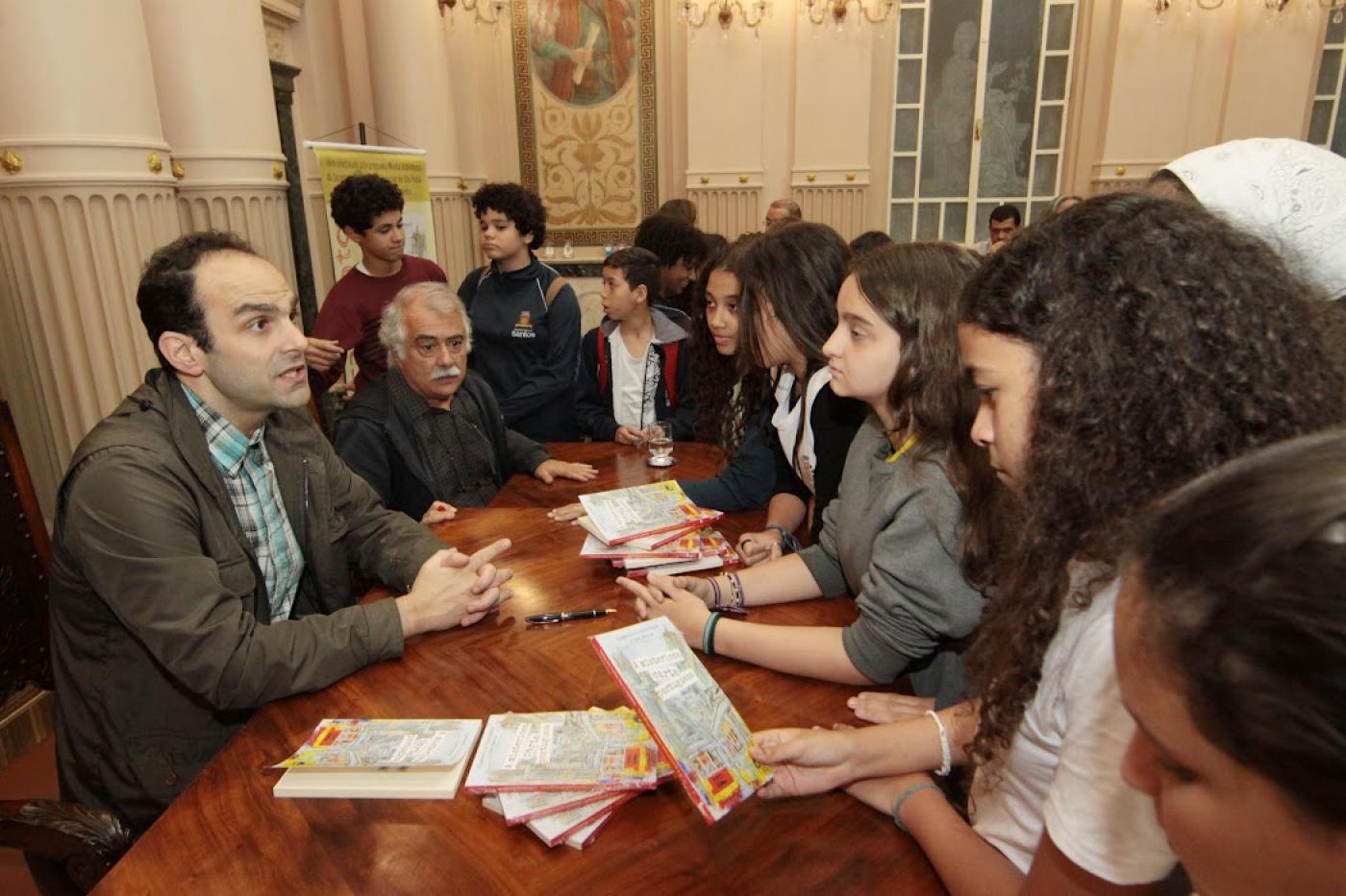 Autores, sentados, conversam com estudantes, em pé, que observam o livro. #paratodosverem