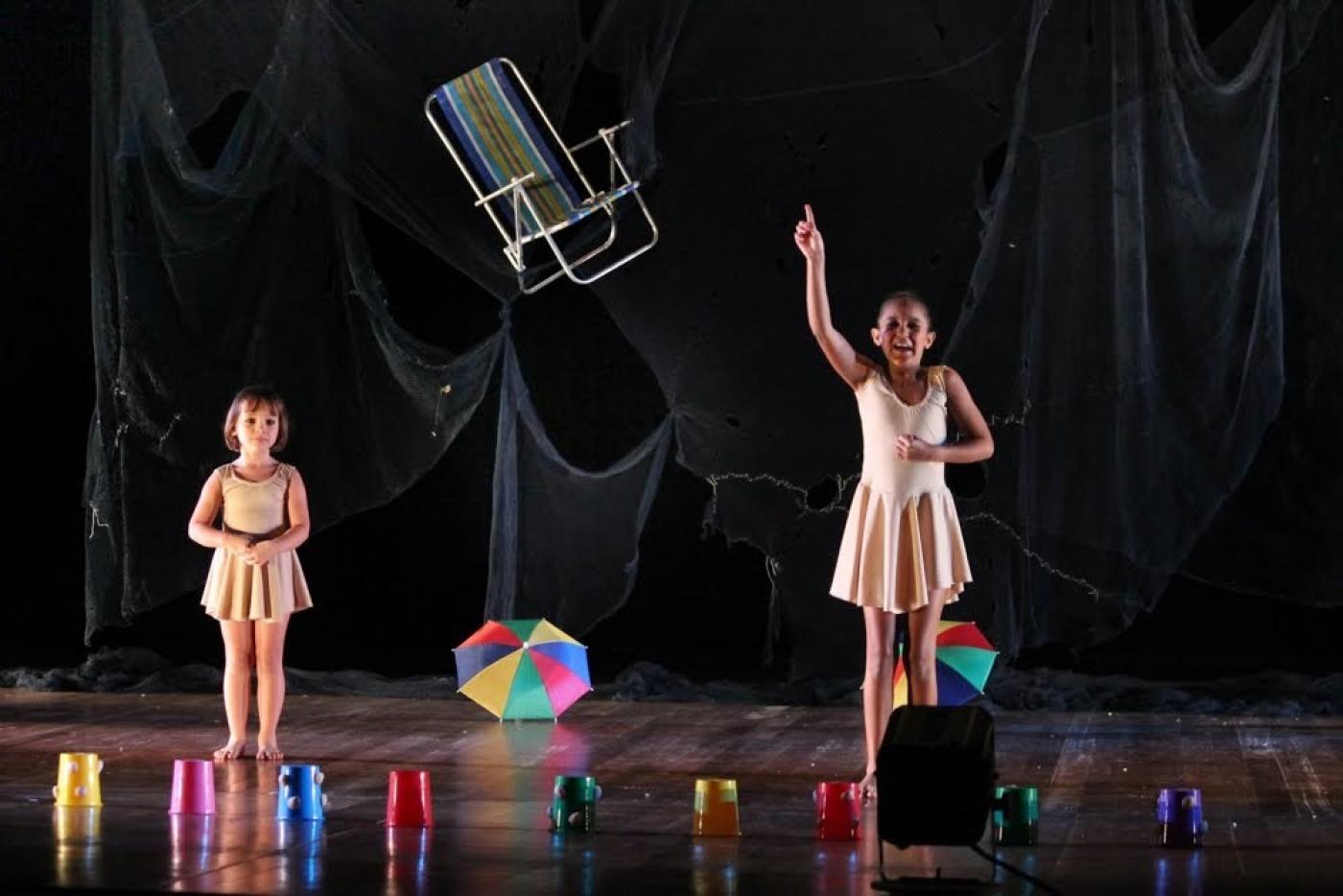 CRIANÇAS APRESENTAM ESPETÁCULO EM PALCO. sÃO DUAS MENINAS. hÁ UMA CADEIRA DE PRAIA PENDURADA. UM PEQUENO GUARDA CHUVA NO CHÃO E ALGUNS OUTROS ELEMENTOS COLORIDOS.#PARATODOSVEREM