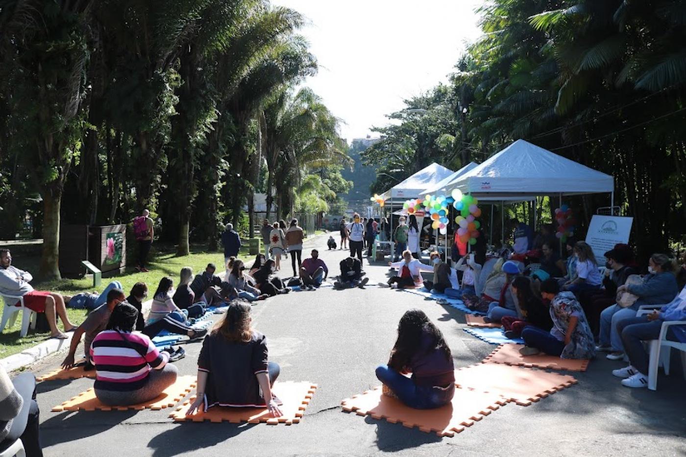 várias pessoas estão sentadas no chão, em roda, participando de uma dinâmica. Há tendas armadas no parque decoradas com balões de gás. #paratodosverem