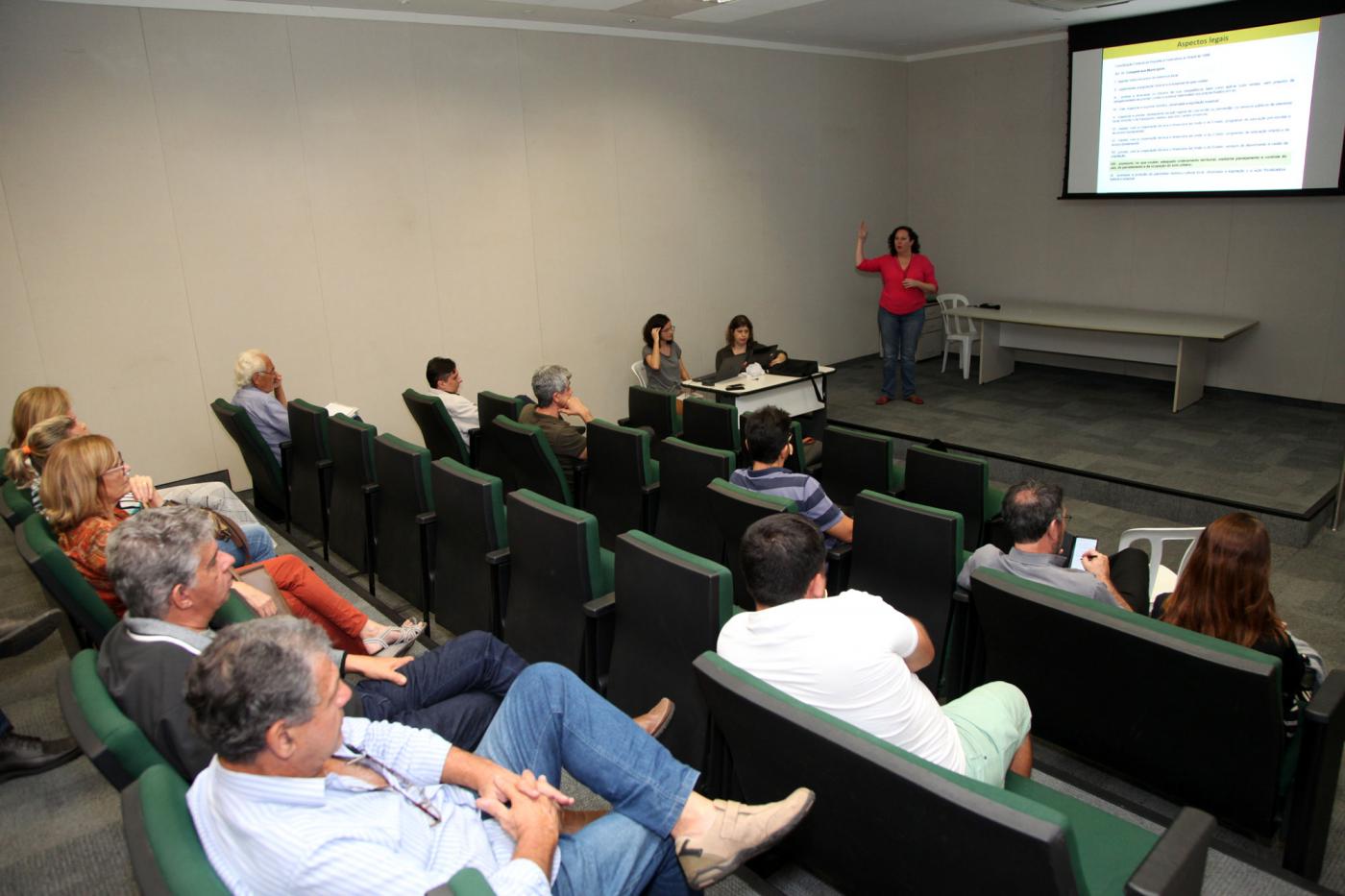 Pessoas estão sentadas em poltronas de auditório. Ao fundo há uma mulher fazendo uma apresentação diante de uma imagem projetada na parede. #Pracegover