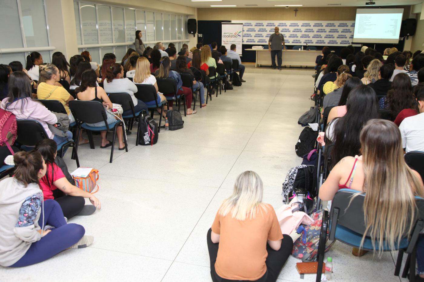 Sala vehia de jovens sentados em cadeiras e até no chão para ouvir palestrante que está ao fundo. #Pracegover