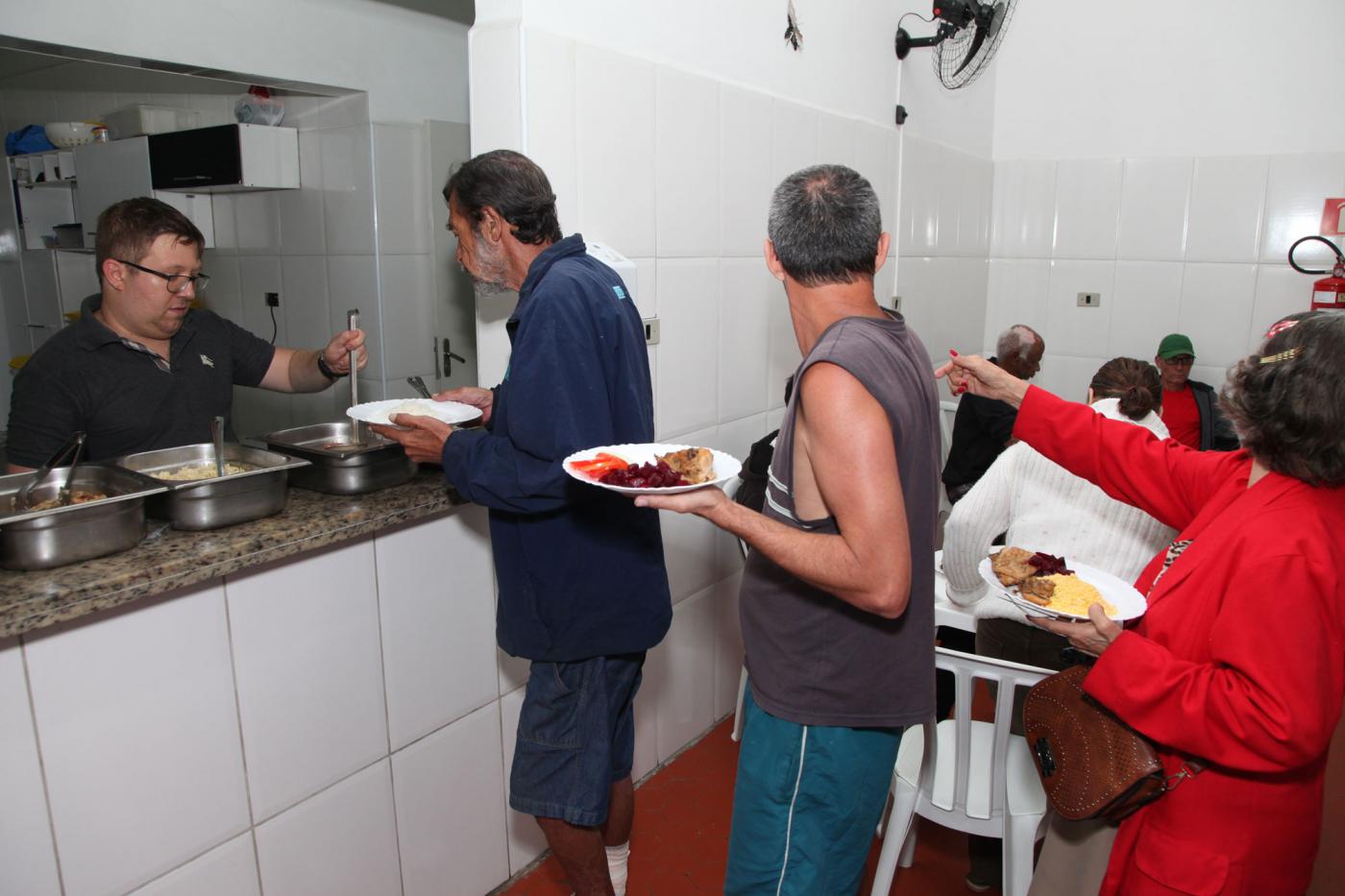 Abrigados em fila para serem servidos. em uma prateleira estão dispostos pratos e uma pessoa server os abrigados. #Pracegover