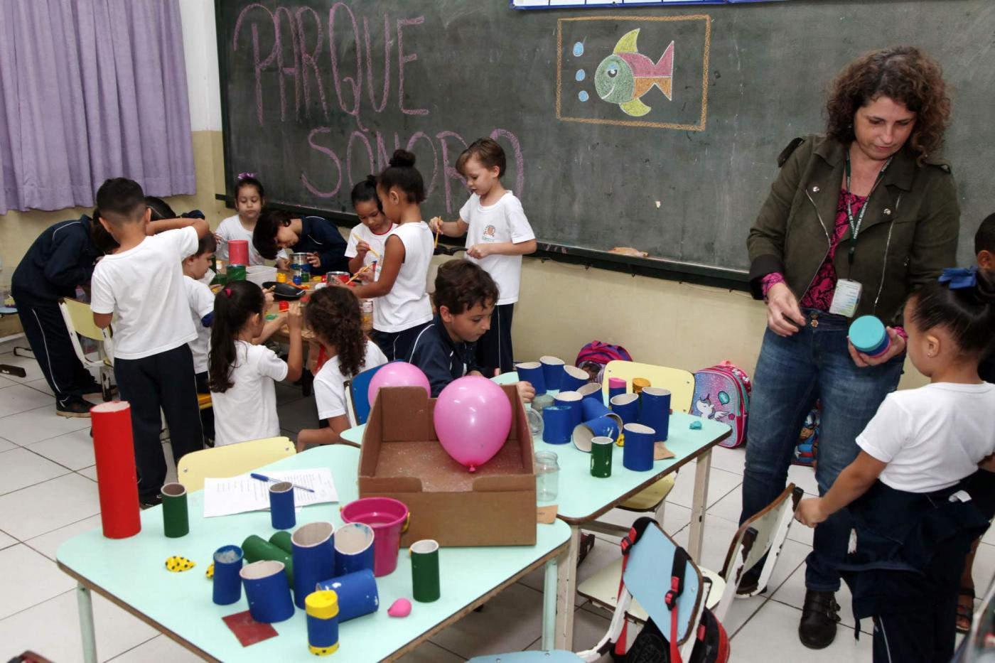 Onze crianças se reúnem em mesa com objetos reciclados. À frente, professora mostra objeto a aluna, junto a mesa com materiais coloridos