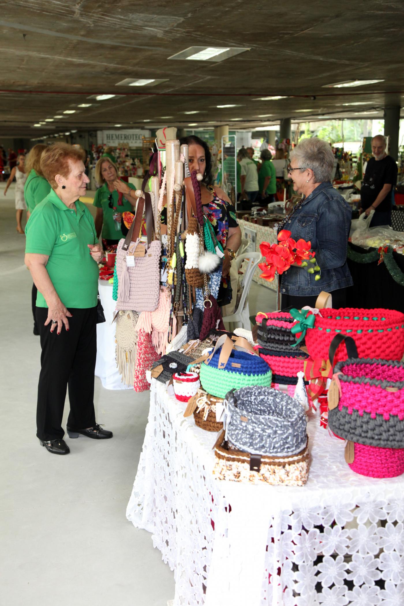 Mesas e suportes cheios de produtos artesanais. Há pessoas circulando pelo local. #Pracegover