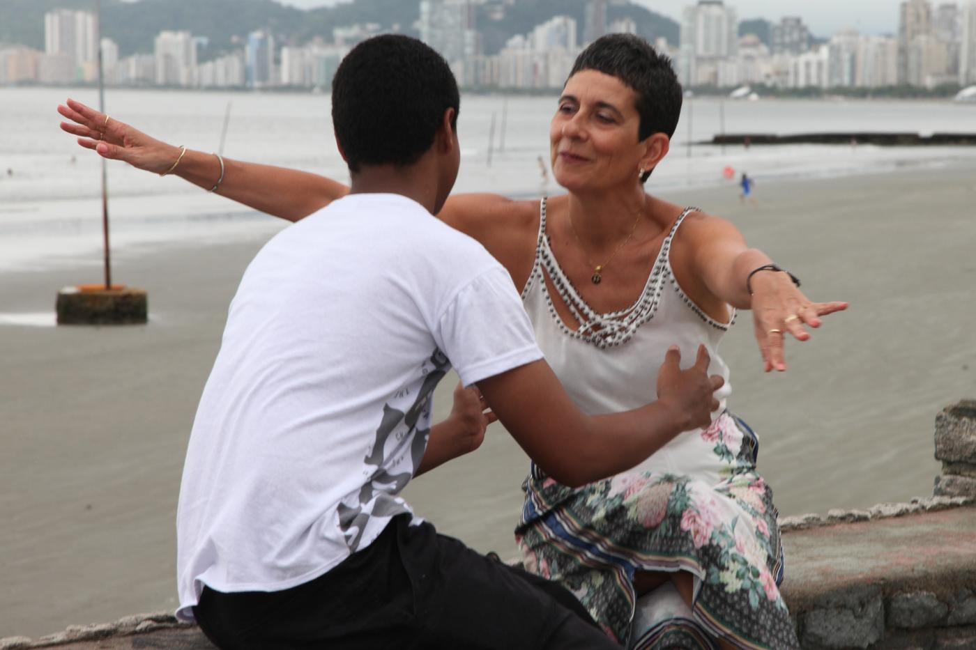 Mulher e jovem se preparam para um abraço. Eles estão sentados em uma mureta de frente para o mar. #Pracegover