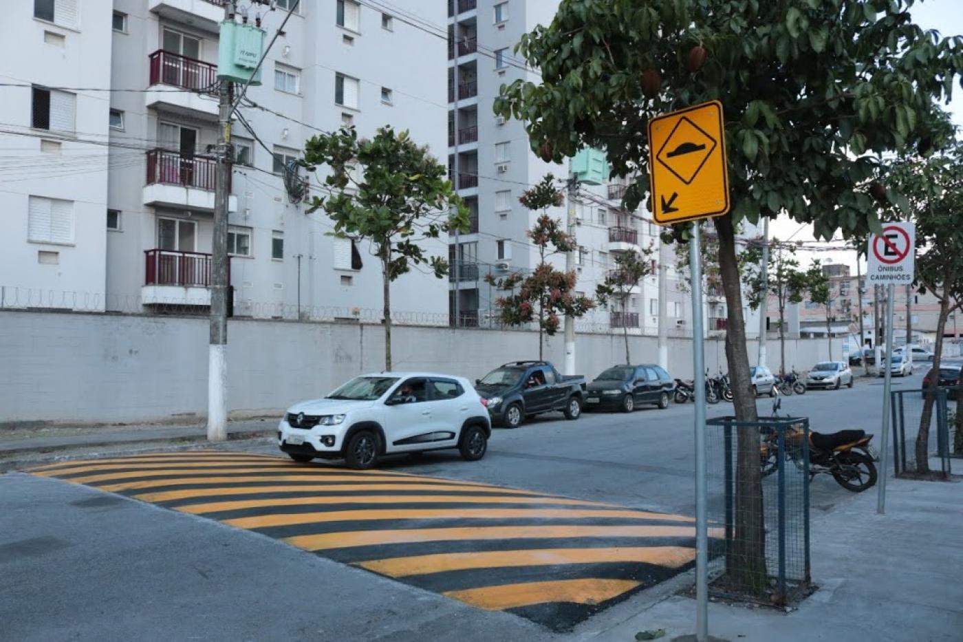 Lombada em primeiro plano com carro branco atravessando em via com prédio de fundo. #paratodosverem