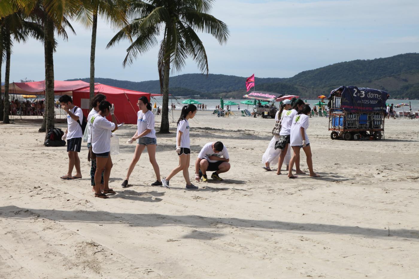 Pessoas caminham na faixa de areia carregando sacos plásticos e recolhendo lixo. #Pracegover