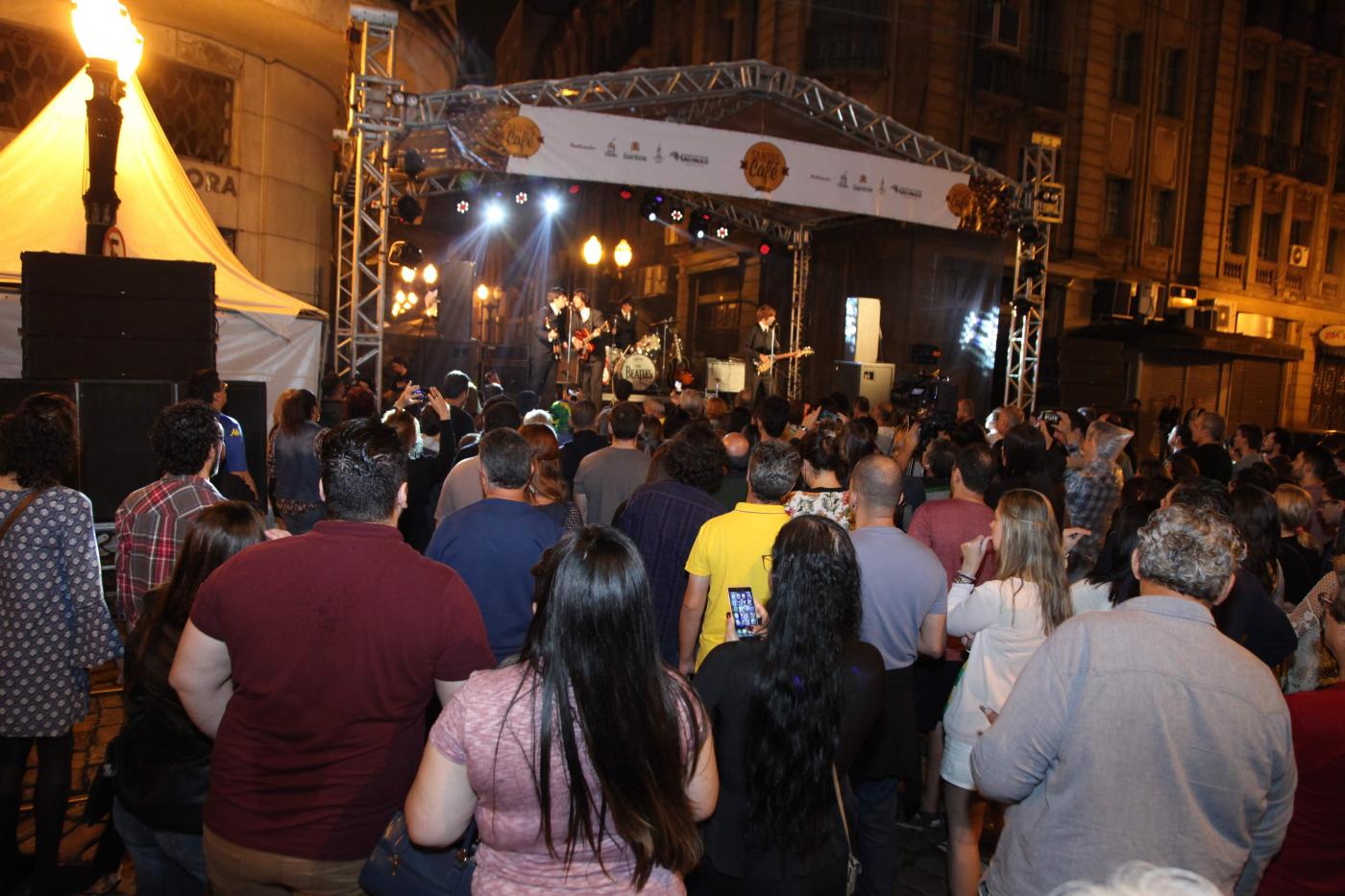 Público está na rua acompanhando show à noite. Ao fundo  há um palco com os músicos. #Pracegover