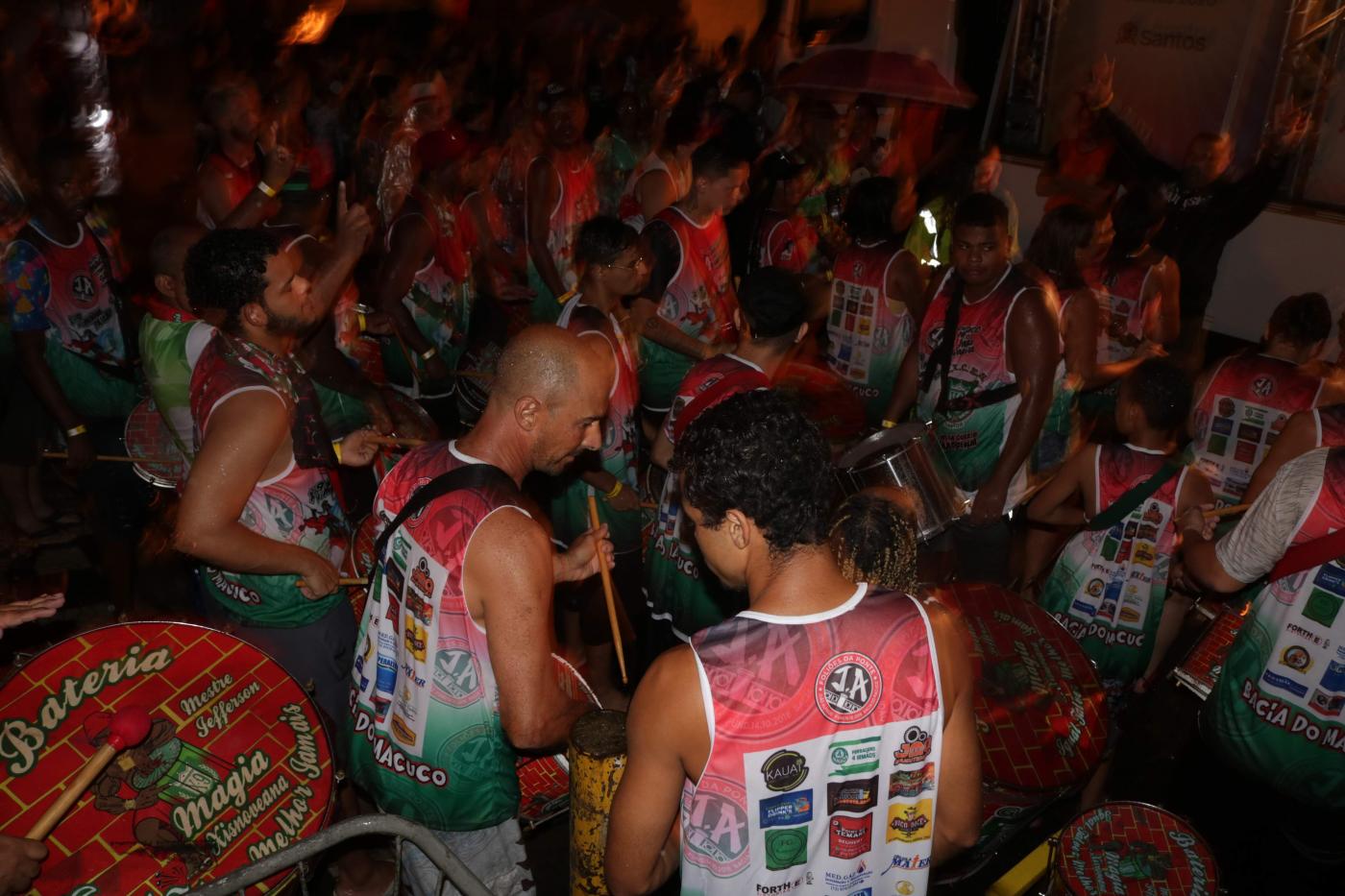 Foliões com camisetas iguais estão em rua com bateria. #Pracegover 