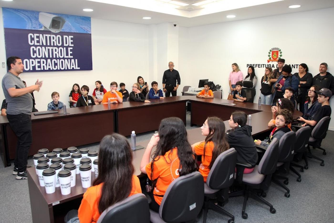 alunos estão sentados em torno de uma mesa ouvido o diretor do CCO. que está em pé. Ao fundo, uma placa onde se lê Centro de Controle Operacional. #paratodosverem