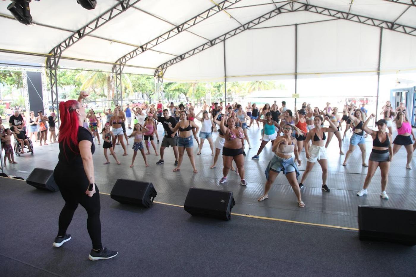 professora dá aula para grande grupo de pessoas, predominantemente mulheres, sob tenda. Ela está no palco, em primeiro plano. O publico se movimente, de frente para a foto, ao fundo. #paratodosverem 