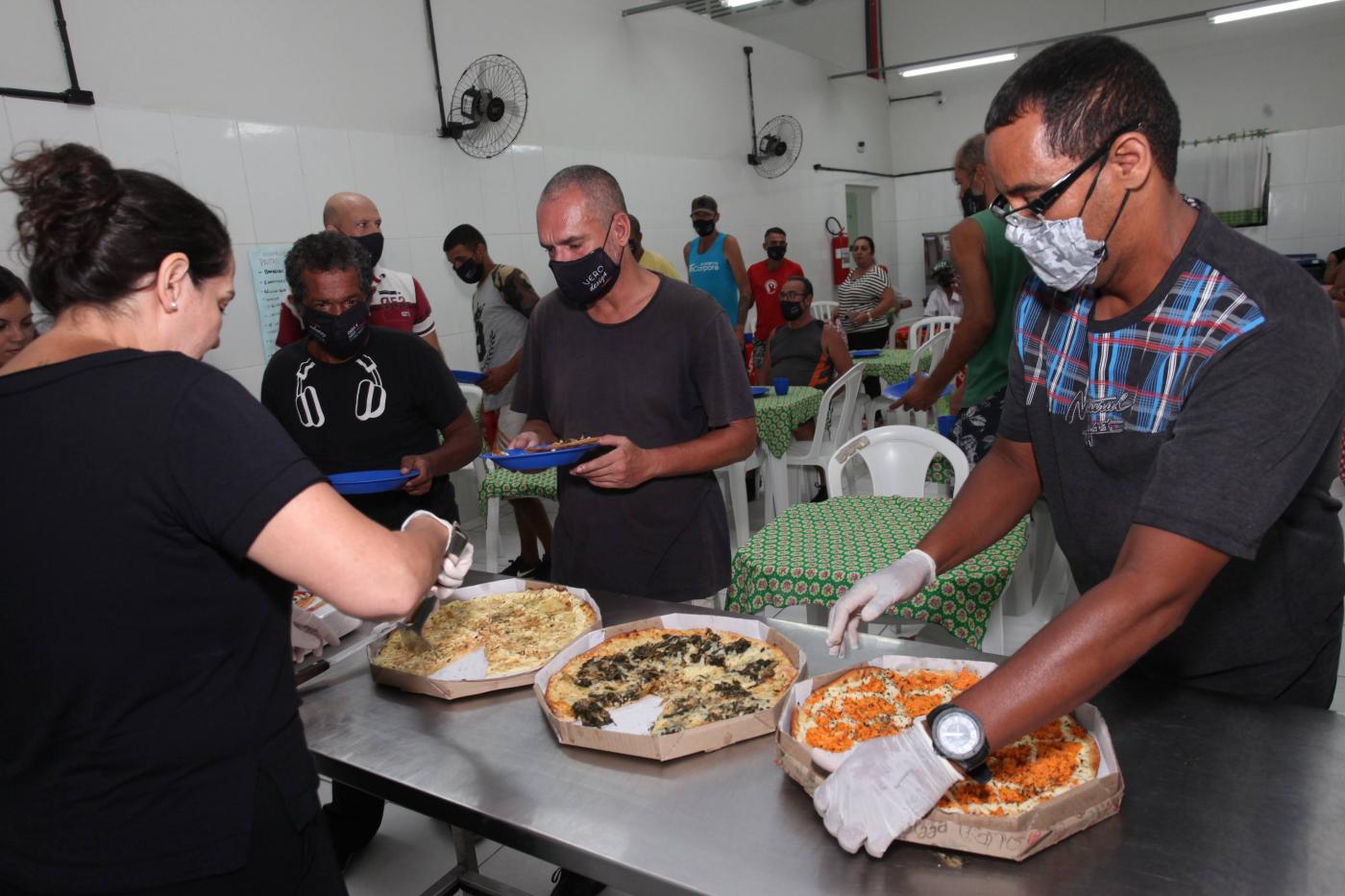 Uma pessoa atrás de uma mesa , onde estão três pizzas , algumas pessoas na fila para comer #paratodosverem