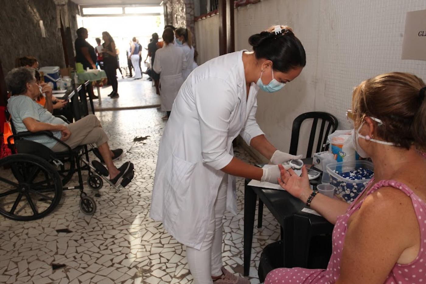 mulher usando avental branco tira gota de sangue da ponta do dedo de outra mulher para teste de glicemia. #paratodosverem