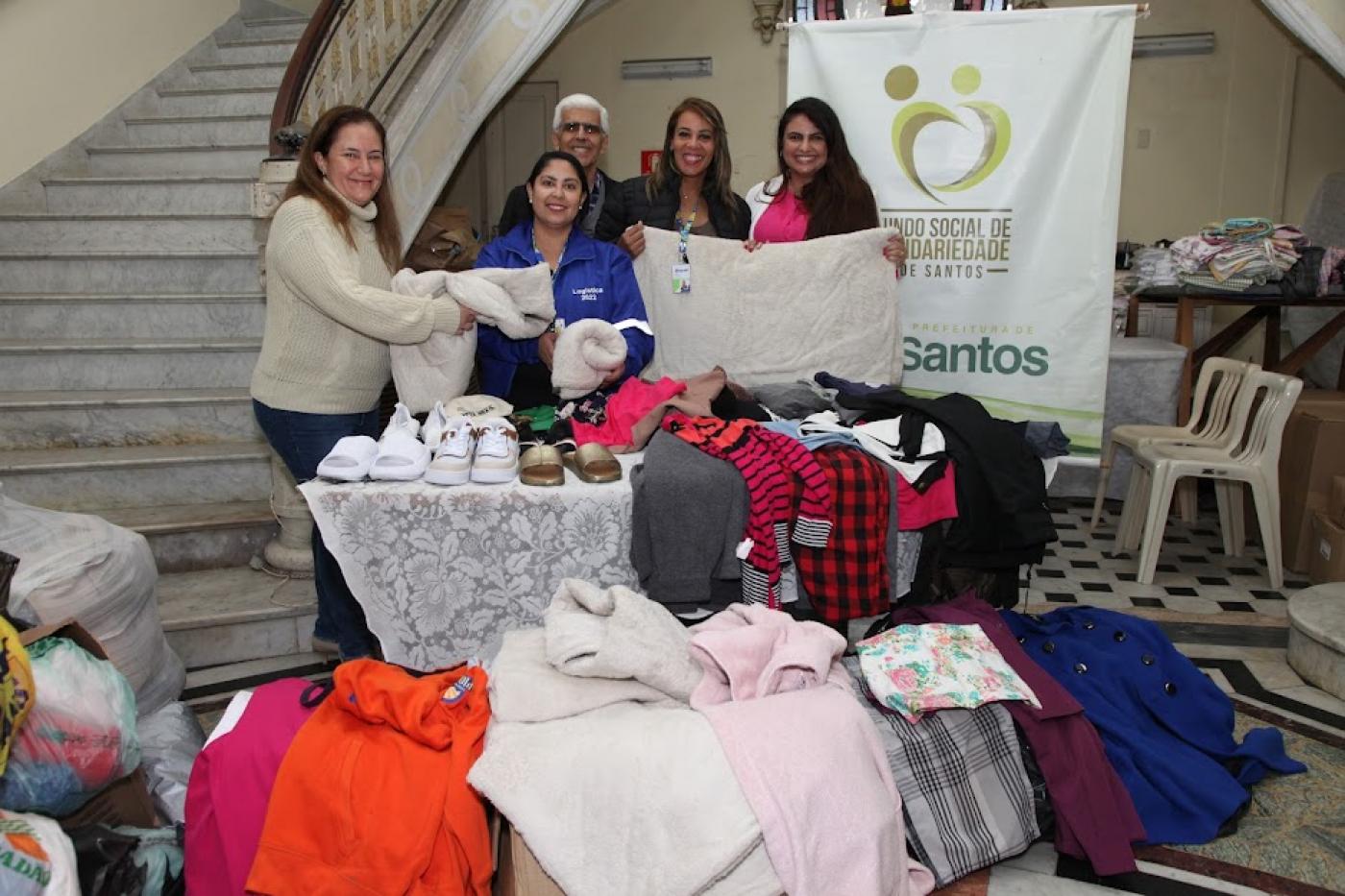 várias roupas sobre caixa e mesas. Mulheres seguram algumas peças e sorriem para foto. #paratodosverem