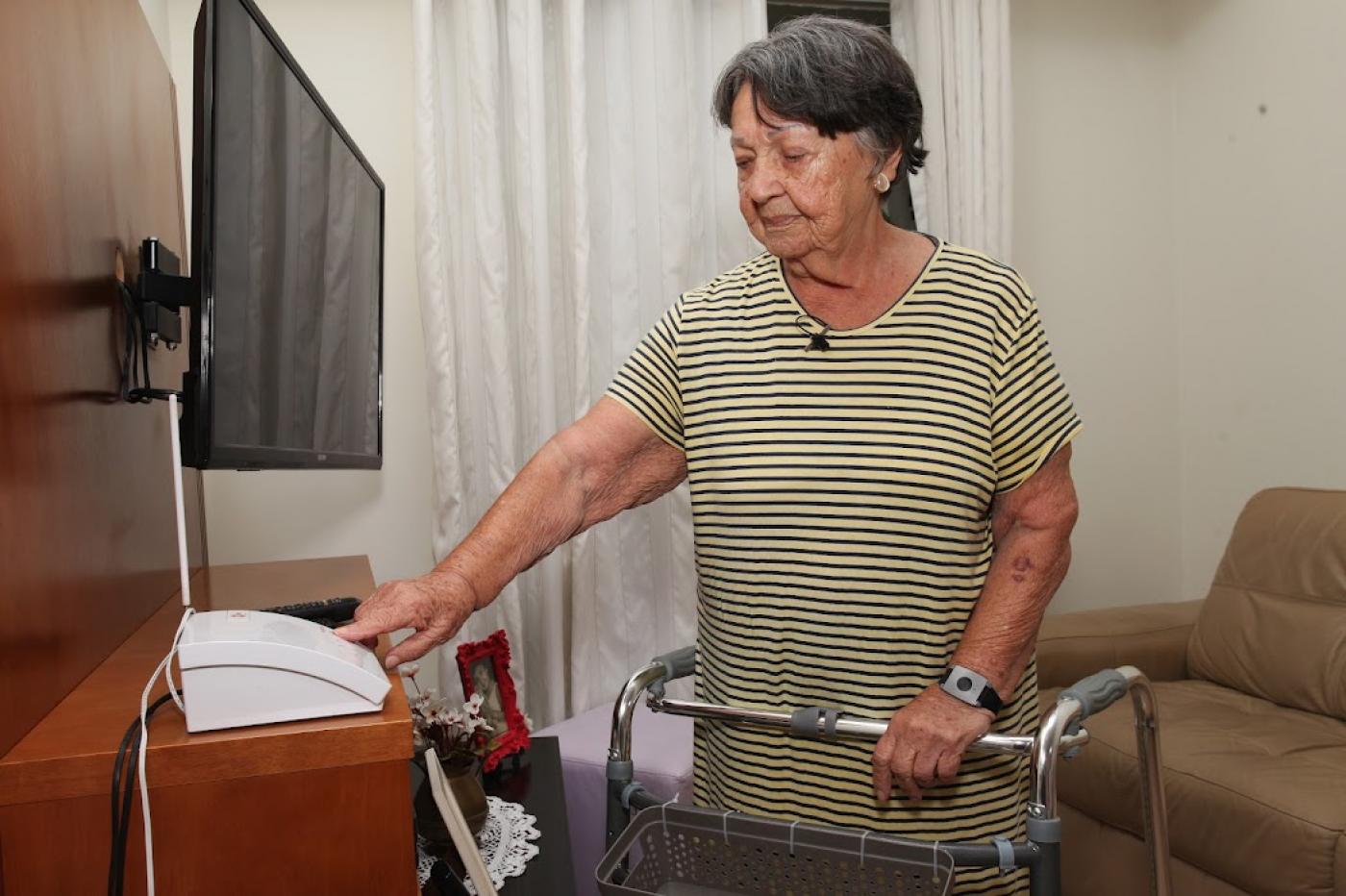 idosa com andador aponta para telefone na sala de seu apartamento #paratodosverem