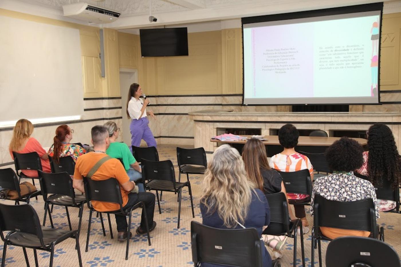 salão com pessoas sentadas assistindo uma palestra. Ao fundo uma mulher em pé fala ao grupo. #paratodosverem