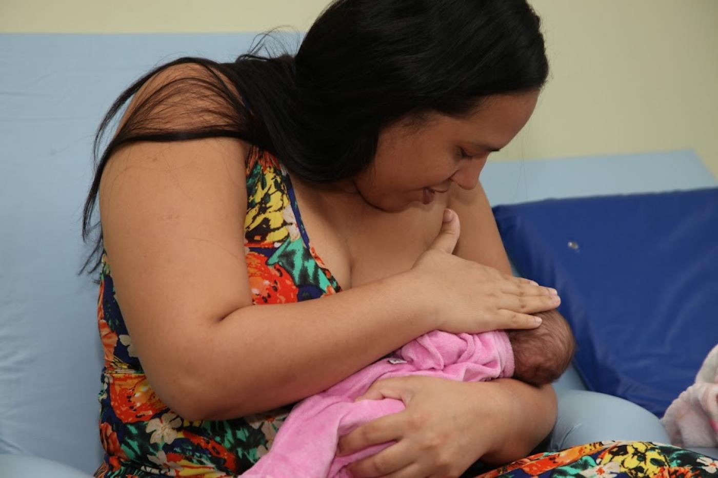 mulher amamenta bebê. Ela cobre o seio e o rostinho da criança ao mesmo tempo em que olha para o bebê. #paratodosverem