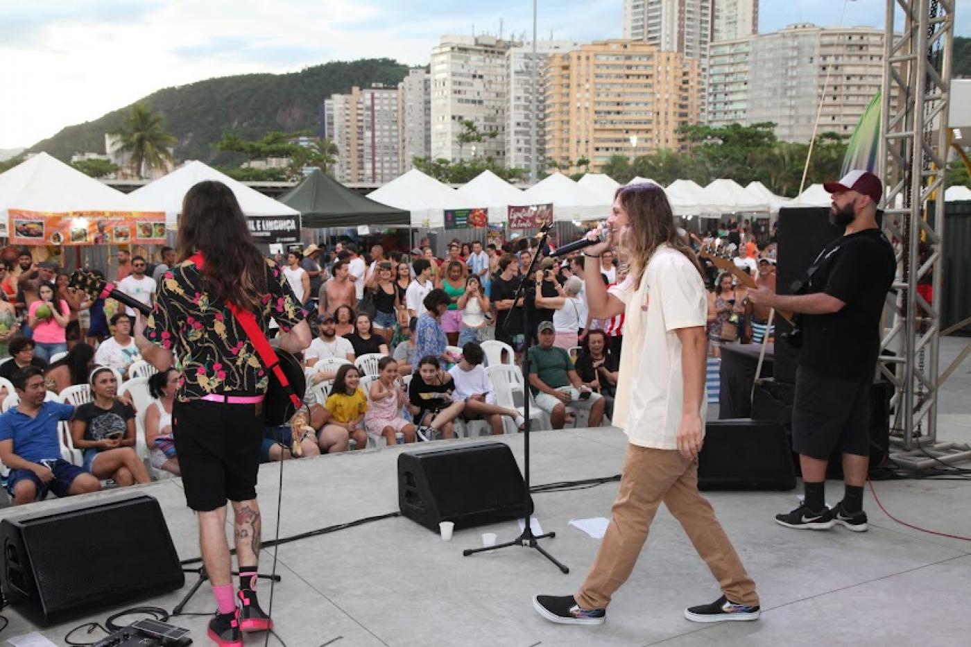 conjunto de três músicos em palco se apresentando para o público ao fundo. #paratodosverem